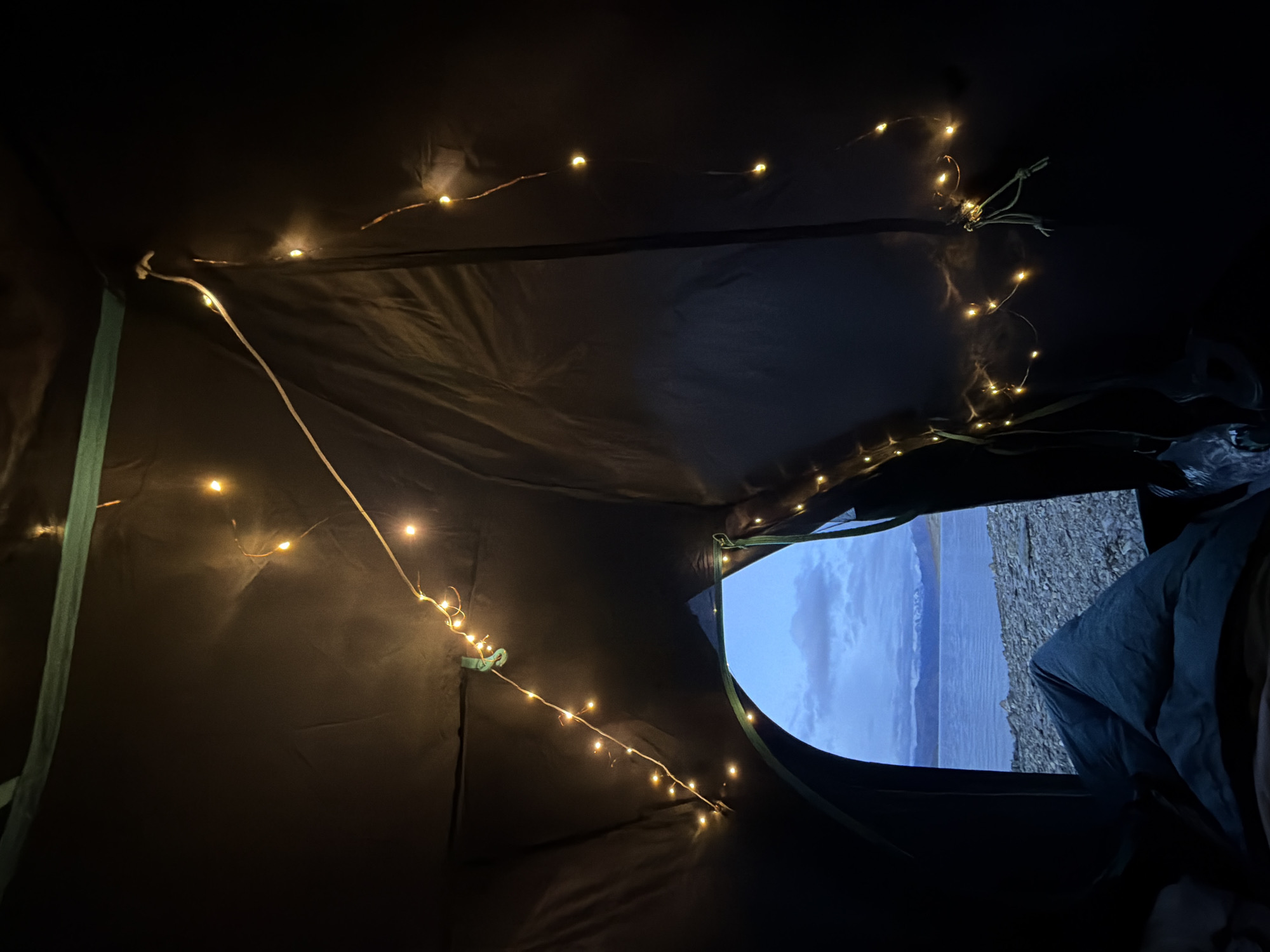 The image depicts the interior view of a dark tent, looking outwards through an open doorway. The tent is decorated with a string of small, warm-toned fairy lights that are draped along the interior walls and ceiling. The lights are illuminated and cast a soft glow.
Through the tent opening, a body of water, likely a lake or large river, is visible. The water appears calm and reflects the dim, dusky sky. Beyond the water, a range of mountains can be seen in the distance. The shore in front of the tent is covered with small, grey stones. A person, only their legs and lower torso visible, is lying down inside the tent, covered by a dark-colored sleeping bag or blanket. The scene suggests a camping or outdoor accommodation setting, possibly in a remote, natural environment. The sky indicates it is either early morning or late evening. The location is described as Unknown, India.