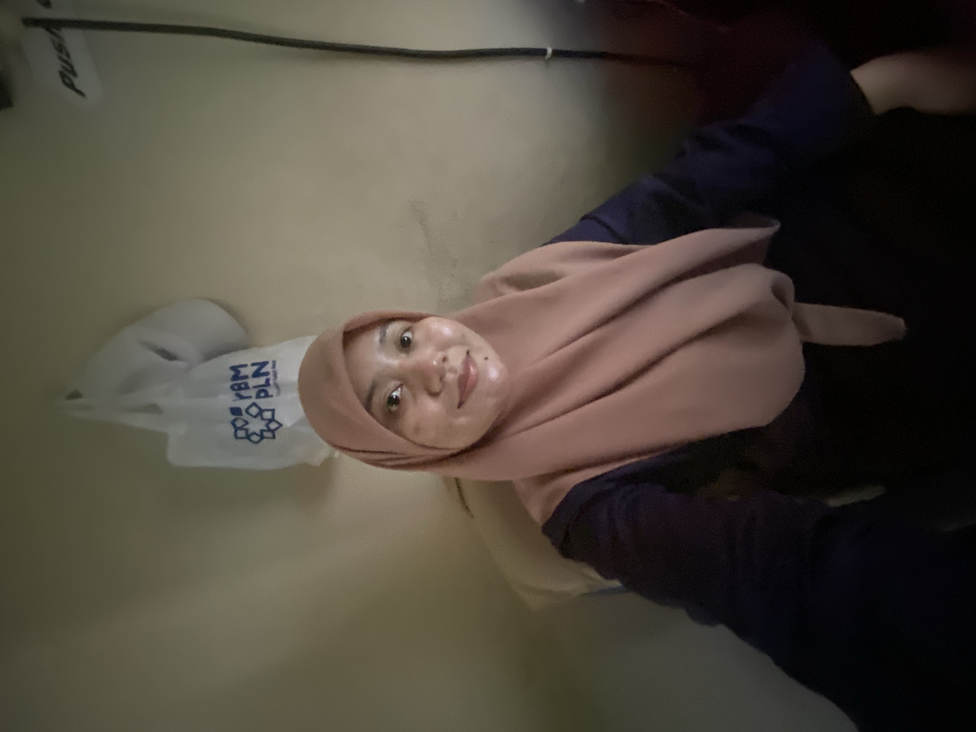 A medium-shot, waist-up, selfie-style photograph depicts a young woman with light brown skin, wearing a mauve hijab and a dark navy long-sleeved top. She is seated, facing forward with a slight smile. The background is a beige wall, with a white plastic bag with blue writing hanging from the wall behind her. A dark grey cable runs vertically down the right side of the image. The lighting is dim. The image appears to have been taken indoors, likely in a residential setting, potentially in the city of Banjarbaru, Indonesia, given the context of the provided information.