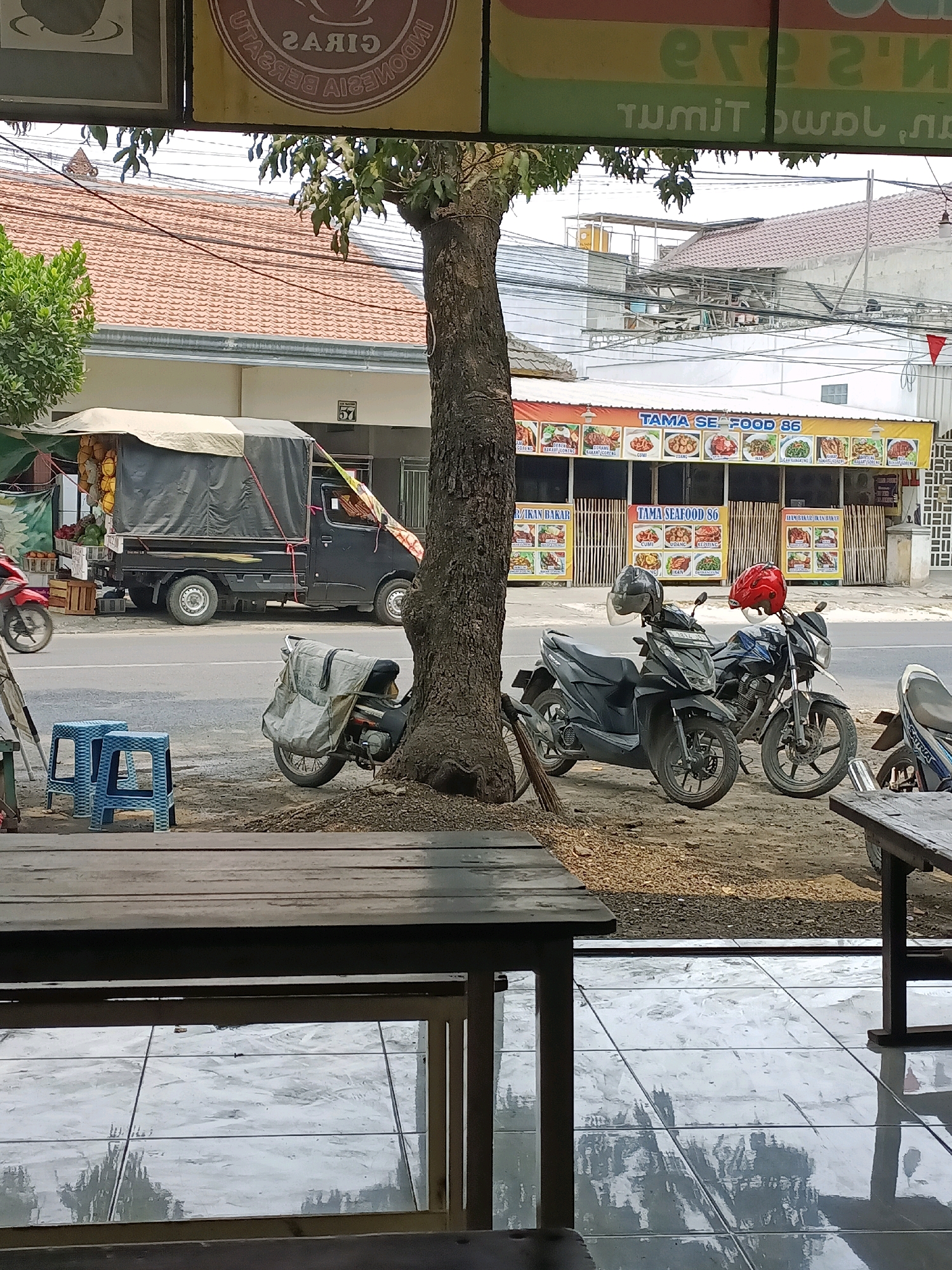 The image captures a street scene in Sukomulyo, Indonesia, likely near a local eatery. In the foreground, a wooden table and chairs are visible, suggesting an outdoor seating area. Beyond this, a tree stands tall, partially obscuring a black pickup truck laden with goods, possibly fruits, covered by a tarp. The truck is parked on the side of a road where a red motorcycle is in motion. Adjacent to the truck, two blue plastic stools are placed on the ground. Further down the road, two motorcycles are parked, one black and one dark blue, both with helmets resting on their handlebars. Behind the motorcycles, a building with the signage "TAMA SEAFOOD 86" is prominent, displaying a menu with various seafood dishes. The overall environment appears to be a commercial area with a mix of residential and business elements, characterized by overhead power lines and tiled sidewalks.