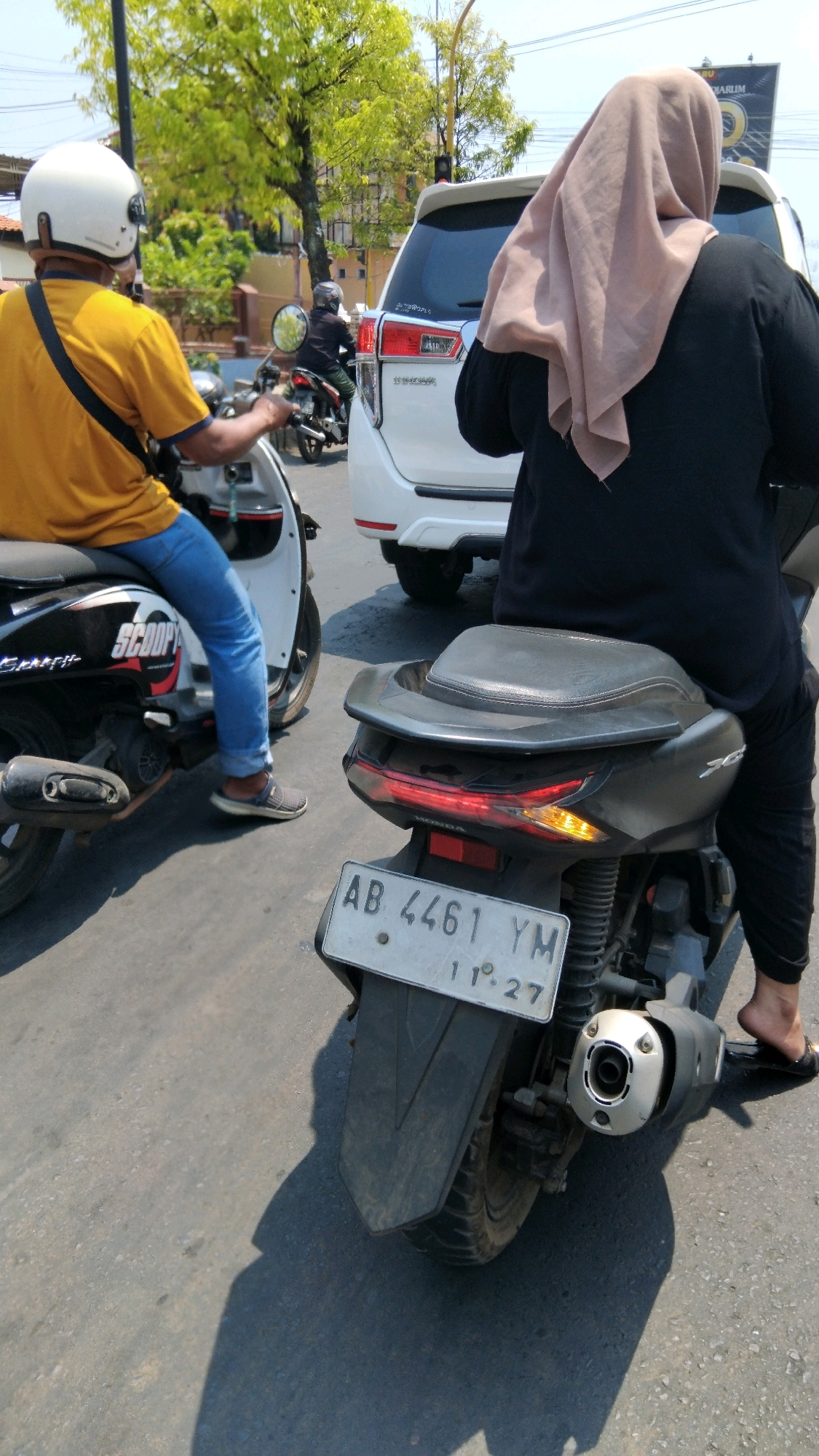 In the city of Batang, Indonesia, on a sunny day, two individuals are seen on motorcycles in traffic. The foreground features a person riding a dark motorcycle, whose legs are clad in black pants, with a tan hijab covering their head and neck. The rear of this motorcycle displays a license plate reading "AB 4461 YM". To the left, another person wearing a yellow shirt and a white helmet is riding a white scooter, identified by the "SCOOPY" logo. This individual is wearing blue jeans and grey shoes. A white SUV is visible behind the person in the hijab, and further in the background, another motorcycle with a rider is passing by. The scene is set on an asphalt road with trees and buildings lining the street.