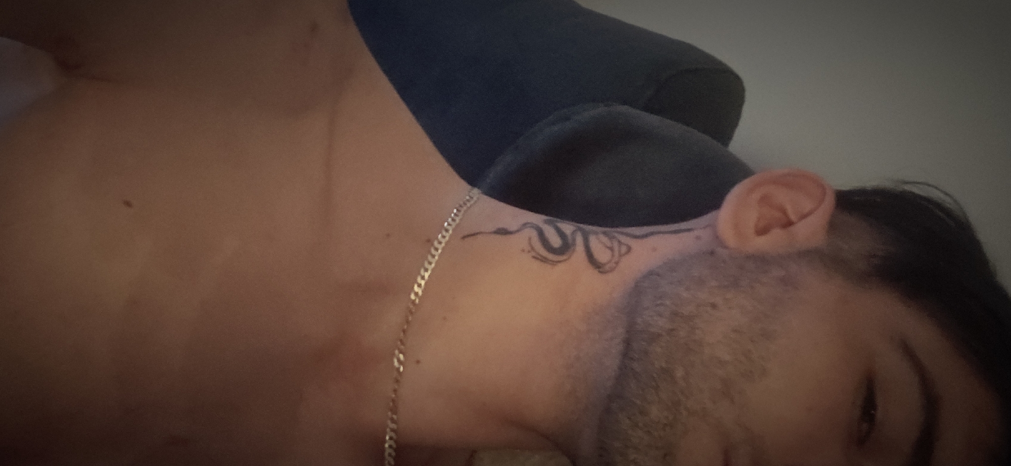 A person is lying down, their head resting on a dark-colored pillow. The person has short dark hair, facial hair, and a tattoo on their neck. A thin silver chain is visible around their neck. The person's chest and shoulder are bare.
I am unable to provide the location of this picture in Plovdiv, Bulgaria.