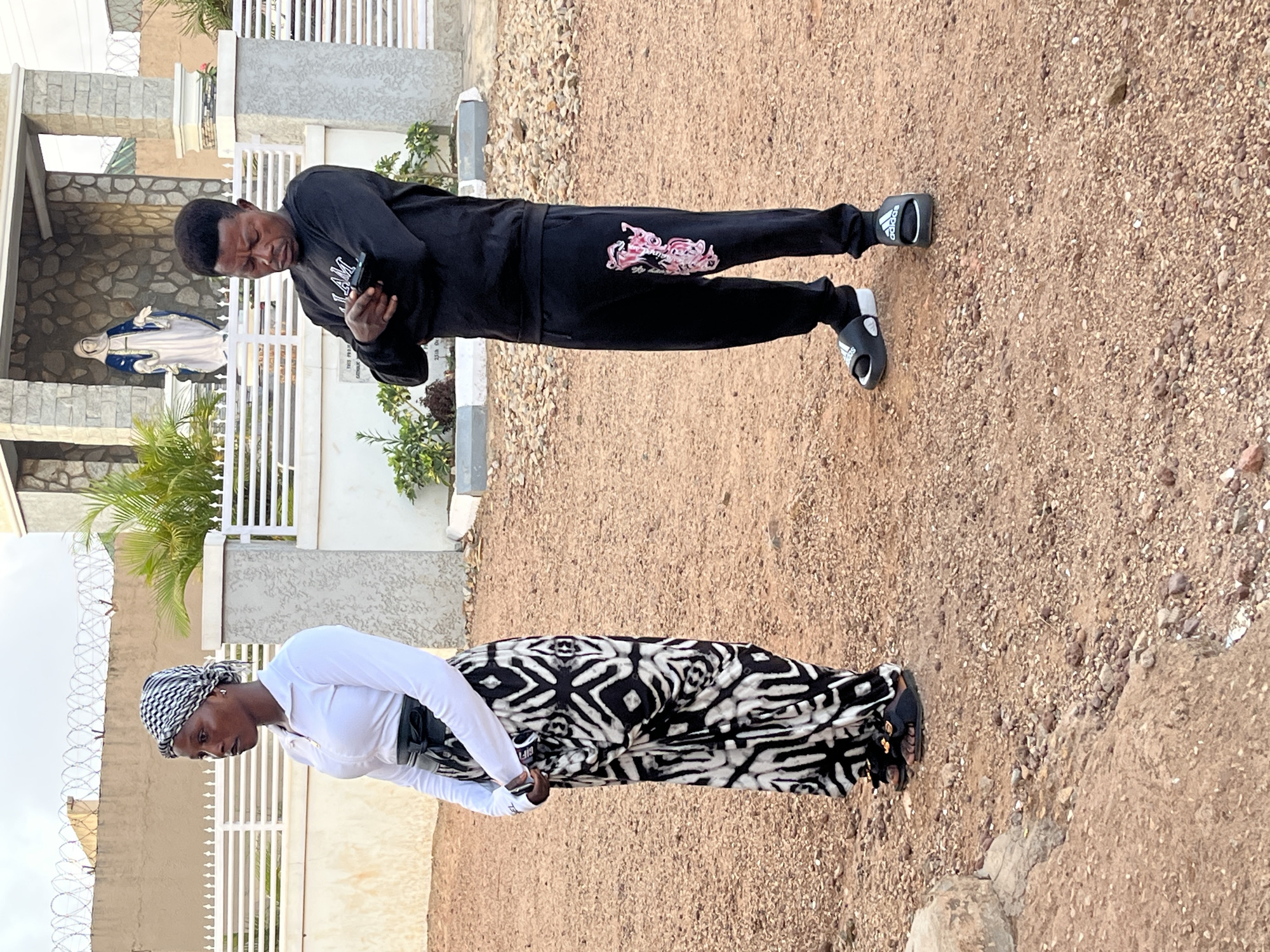 Two individuals stand on a gravel surface, seemingly outdoors. The first individual, a man, is dressed in a black tracksuit with a design on the leg, wearing Adidas slide sandals, and holding a phone. He is positioned near a white structure. Behind him is a statue inside a stone structure with a barred gate. The second individual, a woman, is wearing a white top, a patterned black and white skirt, and a headscarf. She is standing near a wall topped with barbed wire. The environment consists mostly of gravel, with buildings and greenery visible in the background. The location appears to be somewhere in the city of Jos, Nigeria.