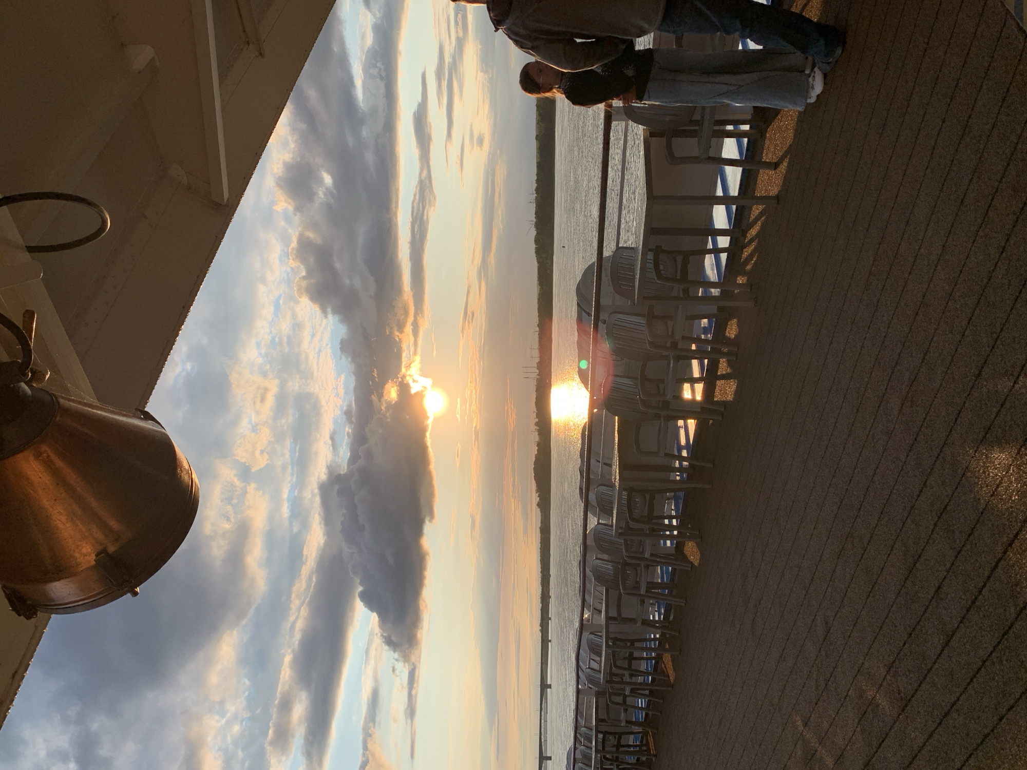 The image captures a sunset scene on a river cruise ship, possibly in the city of Perm, Russia. The sun, low on the horizon, casts a warm golden glow across the water and the sky, which is filled with dramatic clouds. A row of tables and chairs is set up on the ship's deck, indicating an outdoor dining or seating area. Two people are visible on the deck, one a young woman with long hair and glasses, and the other a person from the shoulders down. The foreground shows the wooden deck of the ship, with shadows of the tables and chairs cast by the setting sun. The background features a wide river with a bridge in the distance and a treeline on the opposite bank. The overall atmosphere is serene and picturesque.