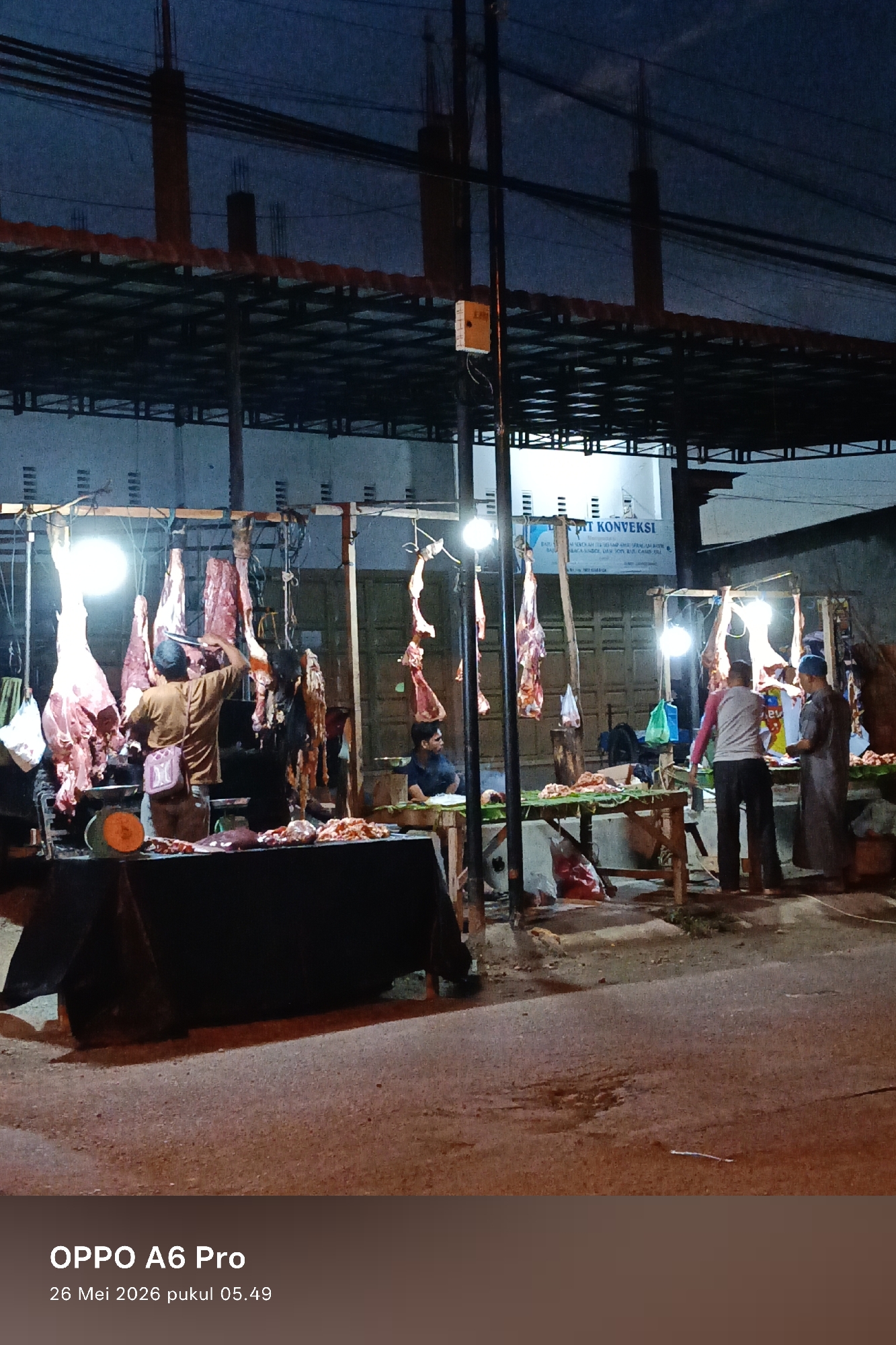 The image captures an outdoor meat market at night. Several carcasses hang from beams under a corrugated metal roof, illuminated by bright lights. Three people stand behind a table, appearing to be vendors, while a customer is present. Meat is displayed on the tables.
The photo was taken in Lhokseumawe, Indonesia, on May 26, 2026, at 5:49 AM.