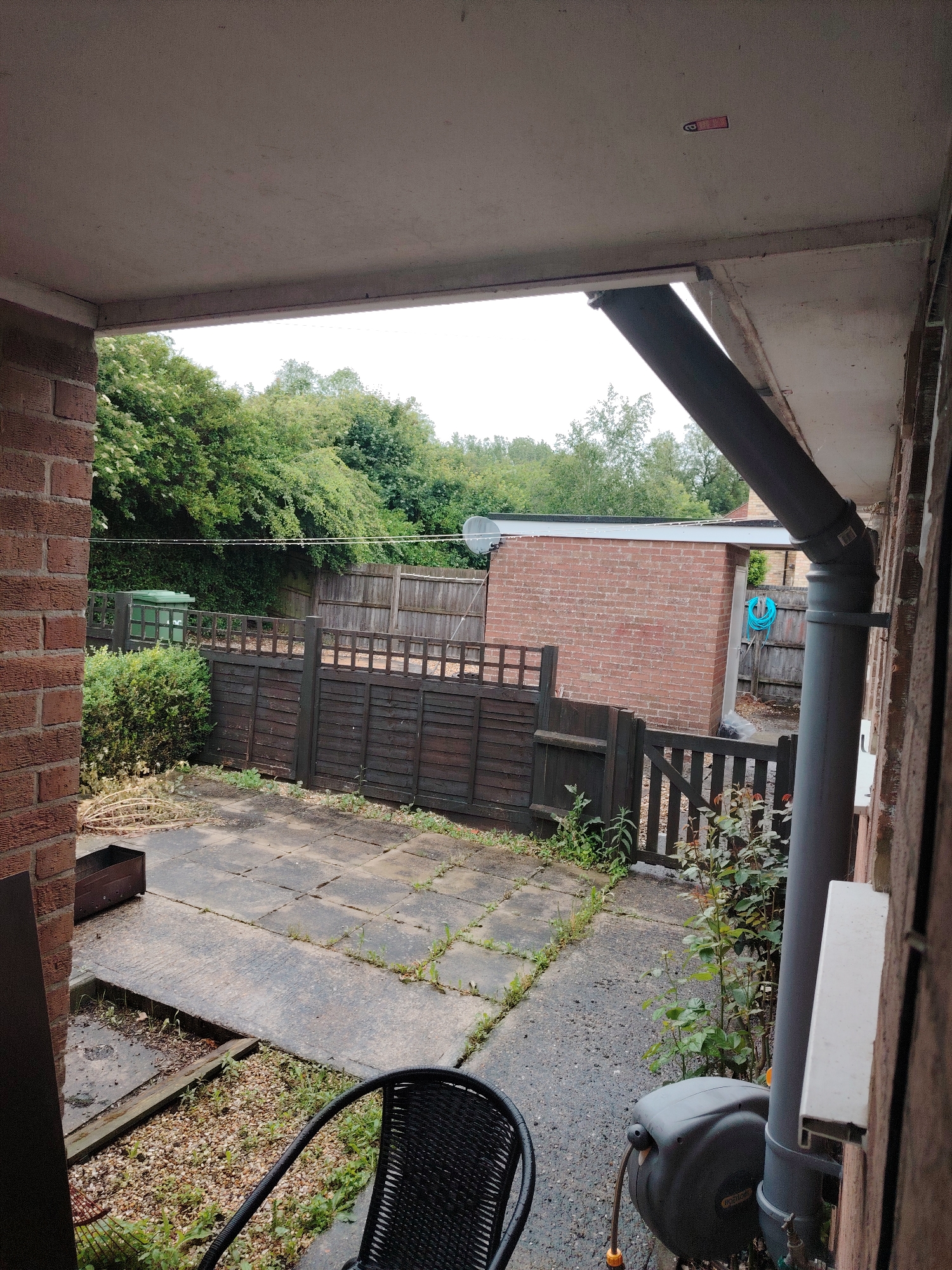 The image depicts an exterior view of a residential backyard or patio area in Huntingdonshire, United Kingdom, observed from under a covered structure on an overcast day.
The overhead ceiling of the covered structure, composed of a light grey, possibly concrete surface, spans the top portion of the image. A small, rectangular red device, featuring a dark symbol (possibly a smoke detector or sensor), is affixed to this ceiling in the upper-right quadrant. A red-brown brick wall is prominent on the left side of the frame, indicating the structure's support. On the right, a grey plastic downpipe descends vertically, secured by two brackets to a similar brick wall. Below the downpipe, a grey, wall-mounted hose reel with an orange hose connector is visible. A white window sill extends horizontally on the right wall, above the hose reel. The armrest of a black wicker-style chair occupies the lower-right corner of the foreground.
The ground immediately beyond the covered area features a paved patio comprising dark grey, square concrete slabs, with pervasive weed growth between the sections. To the left, a dark, rectangular planter or container rests on the ground, alongside a pile of dried, light-colored plant stalks. A patch of gravel with scattered green weeds is visible in the lower-midground, to the left of the chair.
Across the midground, a dark brown wooden fence, characterized by horizontal planks and a lattice top section, delineates a boundary. A matching wooden gate is integrated into this fence towards the right. Behind the left portion of the fence, a dense, dark green bush is present. Further back, a large green refuse bin, marked with "ADC," stands against another wooden fence line.
In the background, a single-story, red-brick outbuilding or shed with a flat, dark roof and a light-colored fascia is situated beyond the fence. A coiled blue hose hangs on the exterior wall of this outbuilding. A grey satellite dish is mounted on a pole or support structure to the right of the outbuilding. The distant background consists of a dense treeline or overgrown vegetation, dominated by various shades of green foliage. The sky is uniformly light grey and overcast. No human or animal figures are present in the scene.