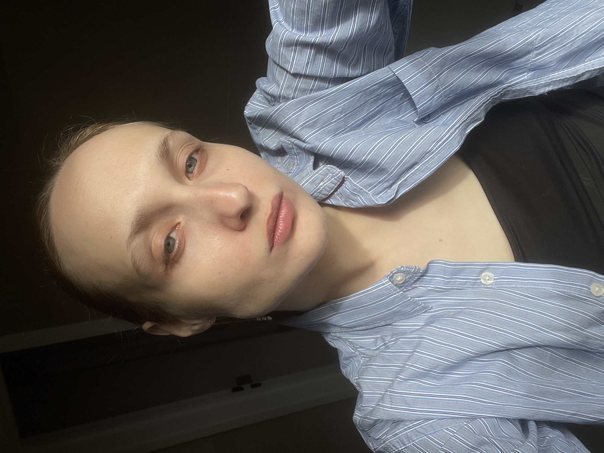 A young woman with light skin and light eyes is the main subject of the image. She has reddish-brown hair pulled back and appears to be wearing a black tank top underneath a blue and white striped button-down shirt. The shirt is partially open. The background is dark and dimly lit, and a portion of a wall or surface is visible in the lower left corner. The picture is taken at an angle. There's no information or evidence to suggest the photo was taken in Подшивалово, Russia.