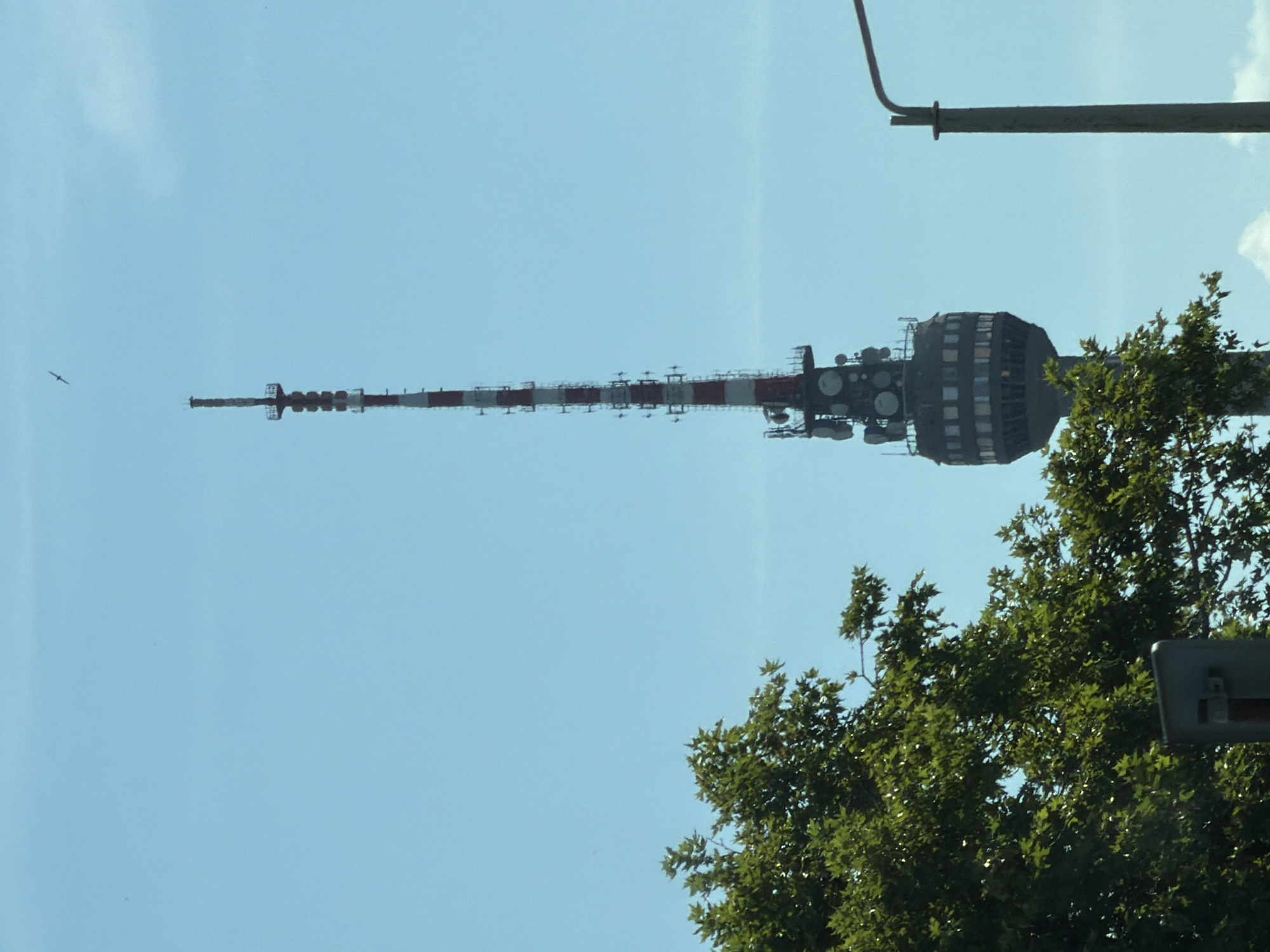The image shows a tall, slender communication tower with red and white horizontal stripes, located in the city of Dolno Ablanovo, Bulgaria. The tower is topped with antennas and a smaller platform. At the base of the tower, there is a circular structure with windows. To the left of the tower, lush green foliage from trees partially obscures the view. A lamppost with a curved arm is visible on the right side of the image, and a bird is flying in the clear blue sky above the tower. The scene appears to be a daytime outdoor shot with a bright sky.
