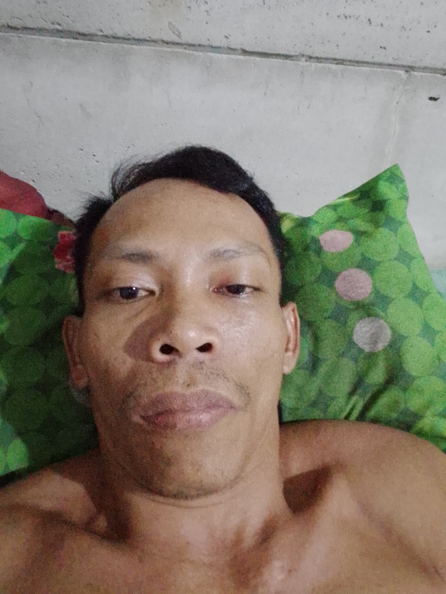 A close-up, eye-level self-portrait of a shirtless Southeast Asian man. He is lying down, possibly on a bed, with a green patterned pillow partially visible behind his head. A white wall is visible in the background. The location is assumed to be Bukit Ketok, Indonesia.