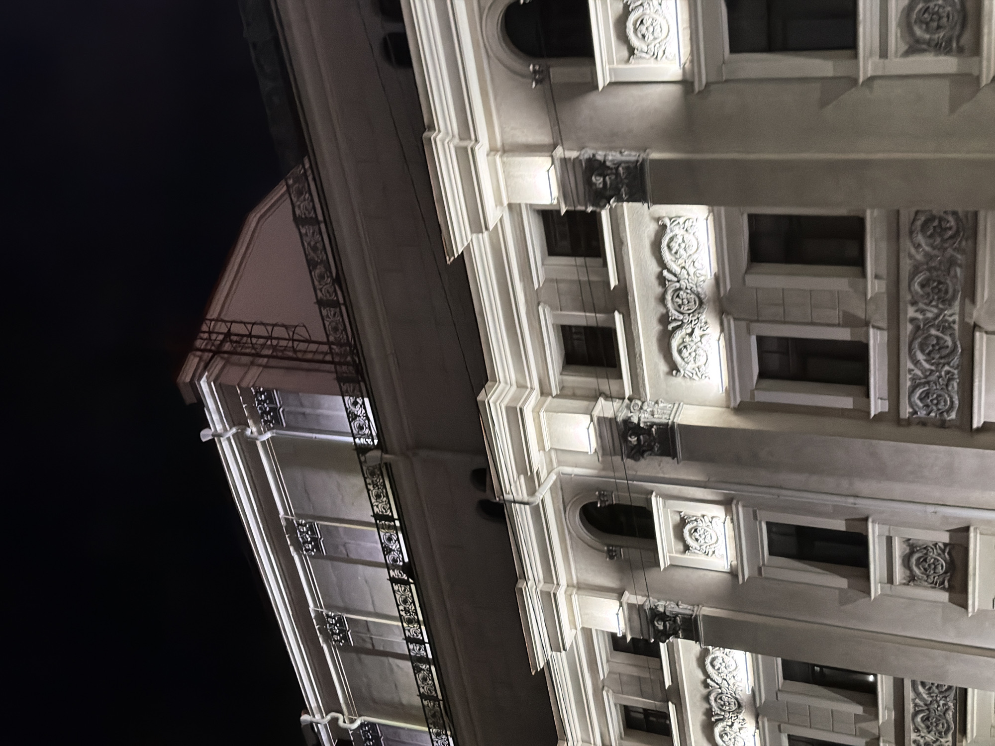 The image shows a low-angle view of a building at night. The building features classical architectural details, including decorative moldings, window frames, and balconies. The building's facade is lit, highlighting its ornate design. The background is a dark night sky. The location cannot be determined from the image.