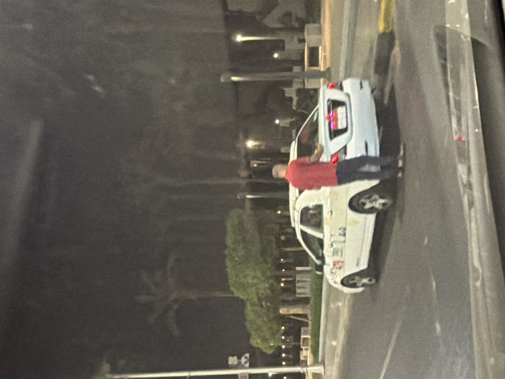 A man in a red shirt and blue jeans is standing next to the rear of a white sedan, possibly a taxi, on a street at night in Sharm El Sheikh, Egypt. The car has markings on its side, including the number "279". Palm trees and streetlights are visible in the background, suggesting an urban or resort setting. The man appears to be engaged in some activity near the car's trunk, though the specific action is unclear. The image is taken from a slightly elevated perspective, likely from another vehicle, as indicated by the blurred foreground.
