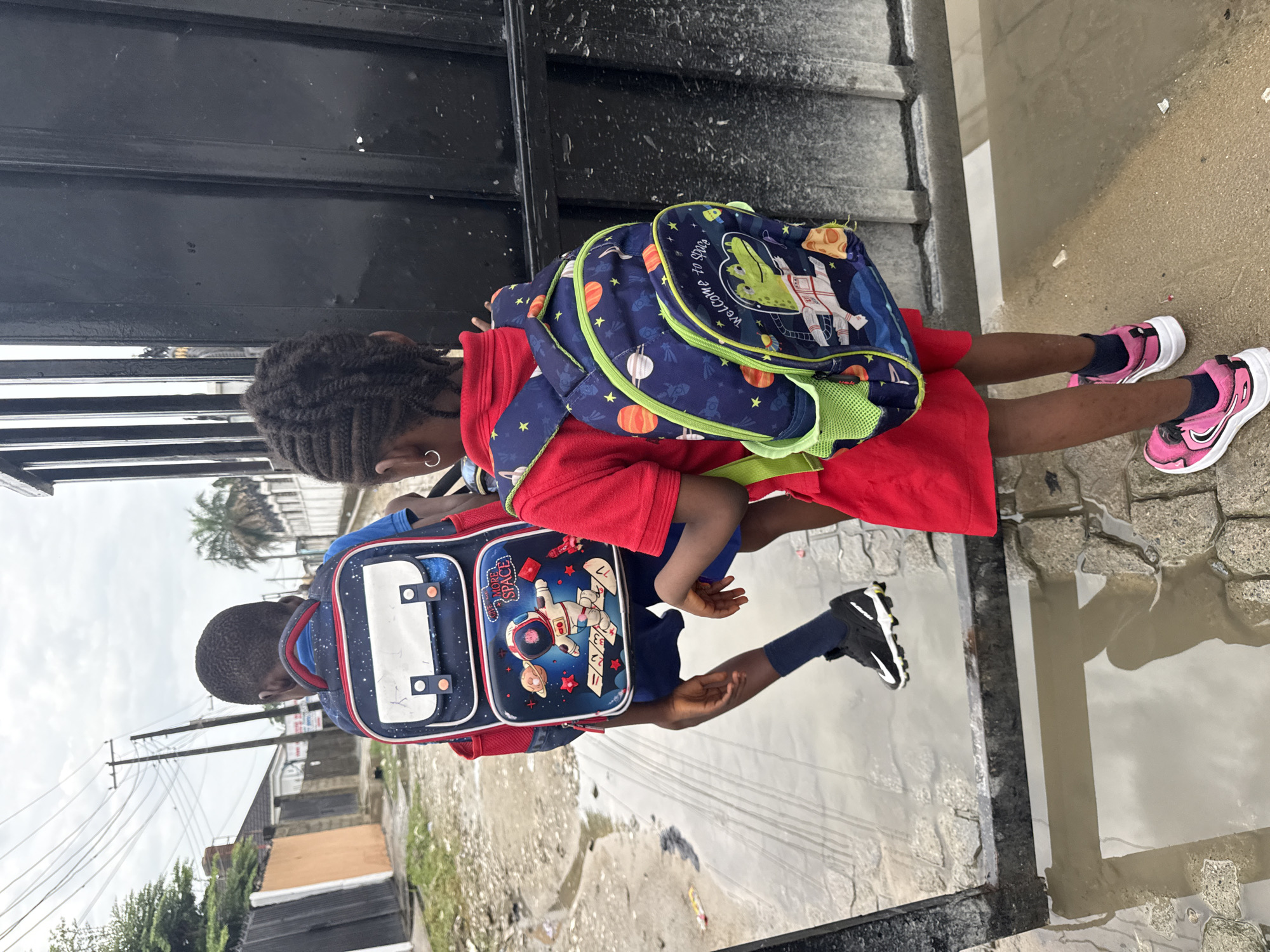 The image, originally rotated 90 degrees clockwise, captures two young children, likely students, walking through a wet outdoor environment.
In the foreground, a boy with short dark hair is seen from behind and to the side. He wears a red top, blue shorts, and black and white sneakers. On his back is a dark blue backpack featuring space-themed graphics, including an astronaut, planets, and rockets. The text "MORE SPACE" is visible on his backpack.
Slightly behind him is a girl with braided dark hair, also viewed from behind and to the side. She wears a red dress or top and pink and white sneakers. Her dark blue backpack is similarly space-themed, showcasing an astronaut, planets, and a distinctive green, crocodile-like alien figure. Visible text on her backpack includes "WELCOME" and partially "Ends at".
The children are navigating a concrete paved area that is significantly wet, with large puddles reflecting the overcast sky. This indicates recent rain or standing water. Adjacent to their path, a dark metal gate or wall runs along the side. The background reveals a residential or semi-urban streetscape with various buildings, numerous utility poles laden with overhead wires, and some sparse vegetation, including a palm tree. The ground in parts appears unpaved or dirtied. The overall setting suggests an established neighborhood, consistent with the provided location context of Ado, Nigeria, under what appears to be a cloudy daytime sky after rainfall. The children seem to be purposefully making their way through the damp conditions, likely to or from school, given their attire and backpacks.