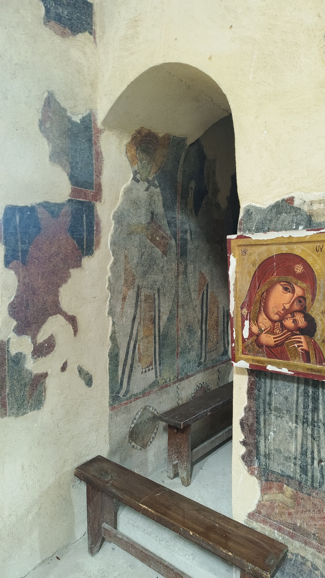 The image shows the interior of a church, featuring deteriorating frescoes on the walls. The paintings are faded and damaged. A framed icon of the Virgin Mary and Child is present on the right wall. Two wooden benches are placed in the foreground. The specific city and country location are unknown based on the image.