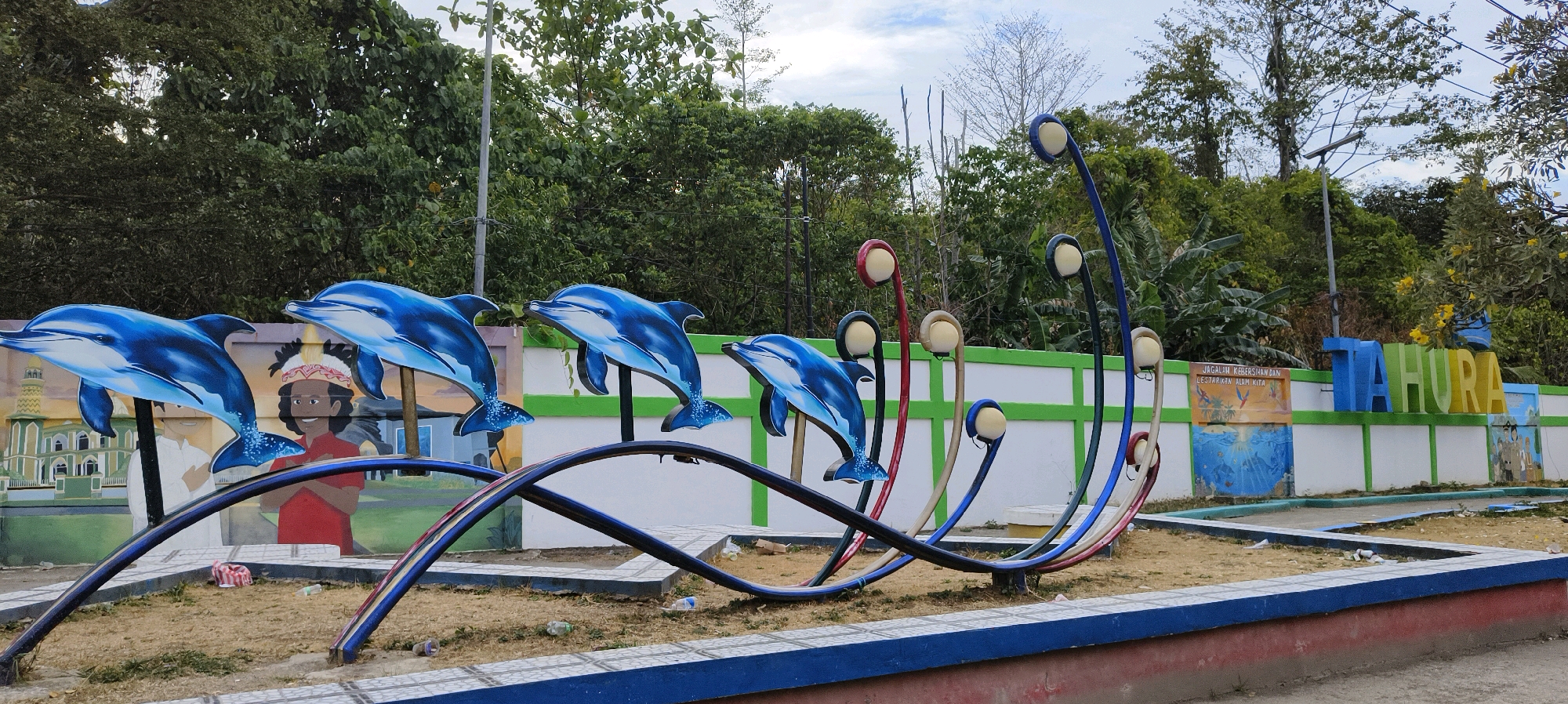In Waisai, Indonesia, a vibrant public space features a playful sculpture of blue dolphins leaping. The dolphins are depicted in mid-air, their bodies arched, with clear, bright blue coloring and white undersides. They are mounted on thin, dark poles. Behind the dolphin sculpture, a mural adorns a white wall with a green border. This mural depicts a mosque and figures, including a child with a traditional headdress. Further along the wall, large, three-dimensional blue and green letters spell out "TAHURA." Scattered on the ground are some plastic bottles and wrappers, indicating the area is a public space that may require cleaning. Lush green trees and foliage form a natural backdrop, with a cloudy sky overhead. The area in front of the wall is a sandy or dirt ground, enclosed by a low blue and red border.