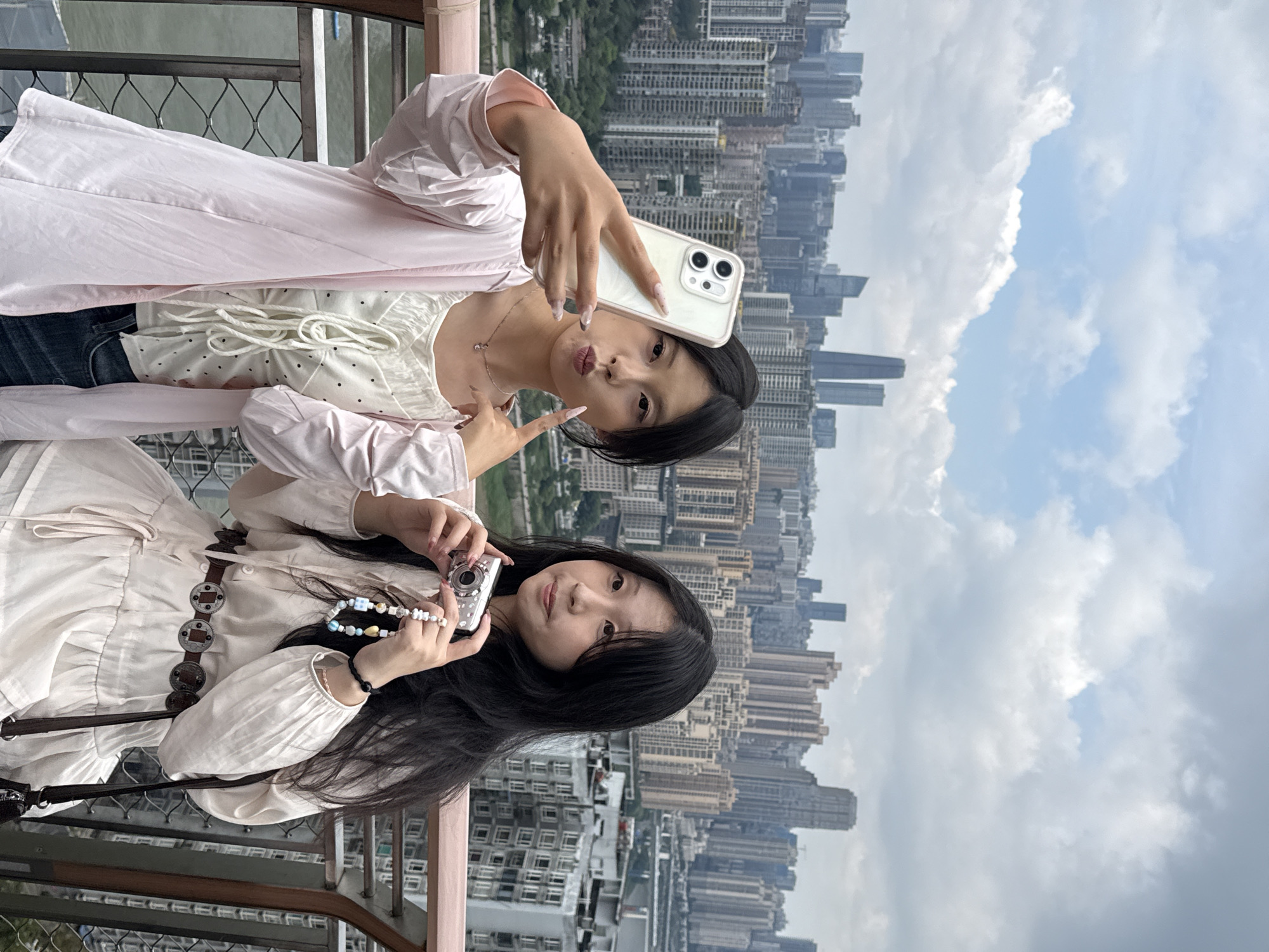 Two young women are taking a selfie in an urban setting. The woman on the top is holding a white smartphone and making a peace sign with her left hand. She is wearing a white top with black dots, a light pink dress, and a necklace. The woman on the bottom is holding a silver digital camera. She is wearing a white dress with a brown belt. The location is Yuzhong District, China, as indicated by the dense cityscape in the background, composed of numerous tall buildings. The sky is cloudy. Both women are facing the camera.