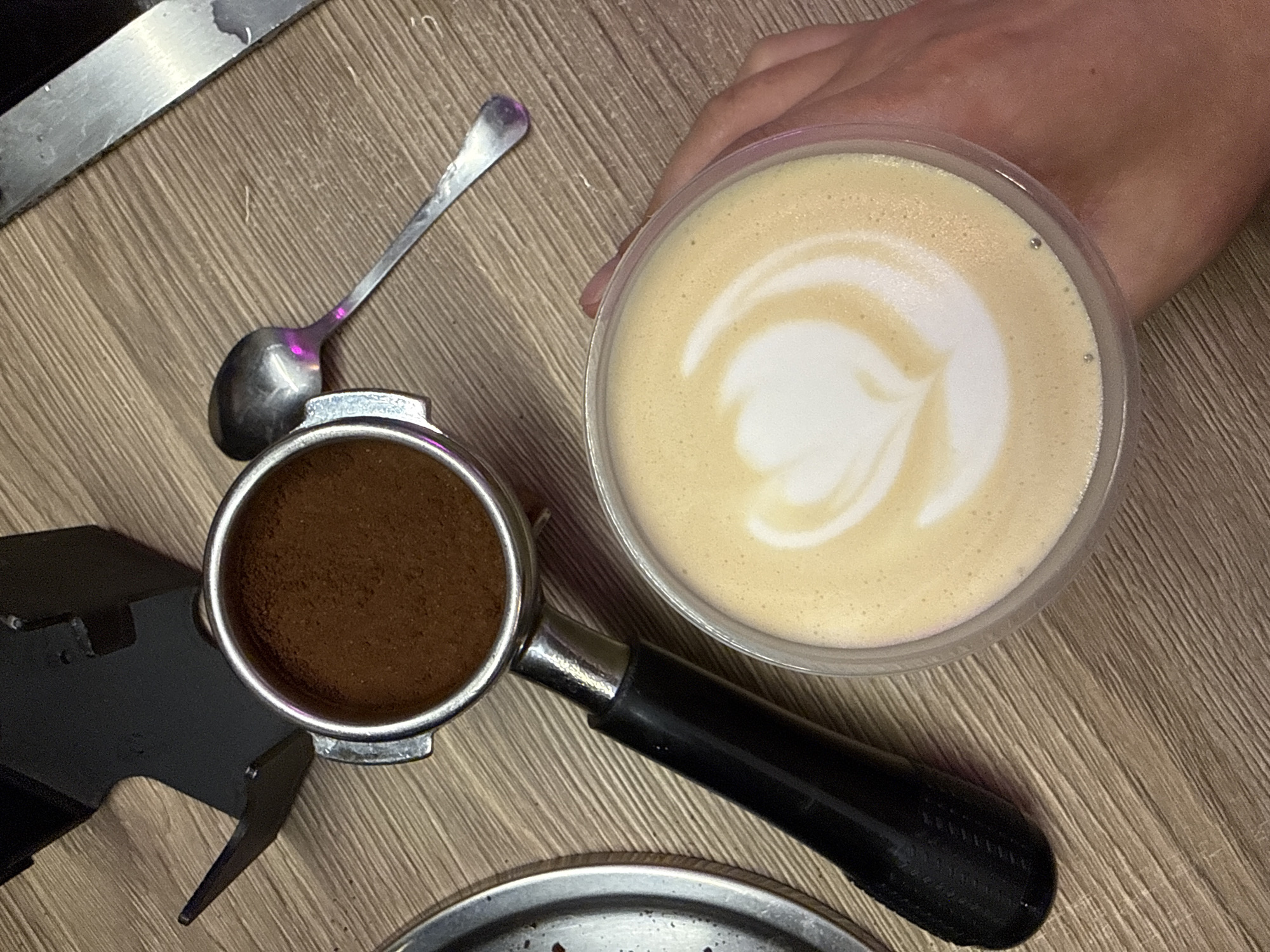 A high-angle shot shows a cappuccino held in a person's hand, an espresso portafilter filled with ground coffee, and a spoon on a wooden surface. The cappuccino is in a clear plastic cup, showing latte art in the foam. The portafilter is stainless steel with a black handle and sits on a metal tamping stand. The spoon is small and appears slightly tarnished. The wooden surface has a light brown color and subtle grain. The location cannot be determined from this image alone. There is no verifiable evidence to show the location is Moscow, Russia.