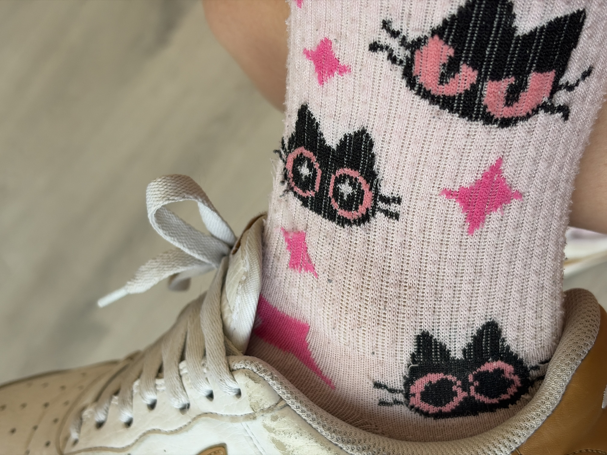 In a close-up, eye-level shot, a person's left foot wearing a white athletic shoe with a tan sole and a pink sock with black butterfly patterns and pink stars is visible. The shoe laces are tied and slightly frayed. The sock is pulled up to just below the knee, and the texture of the knitted fabric is apparent. The background consists of a blurred wooden floor. The image does not provide any information about the location in a city.