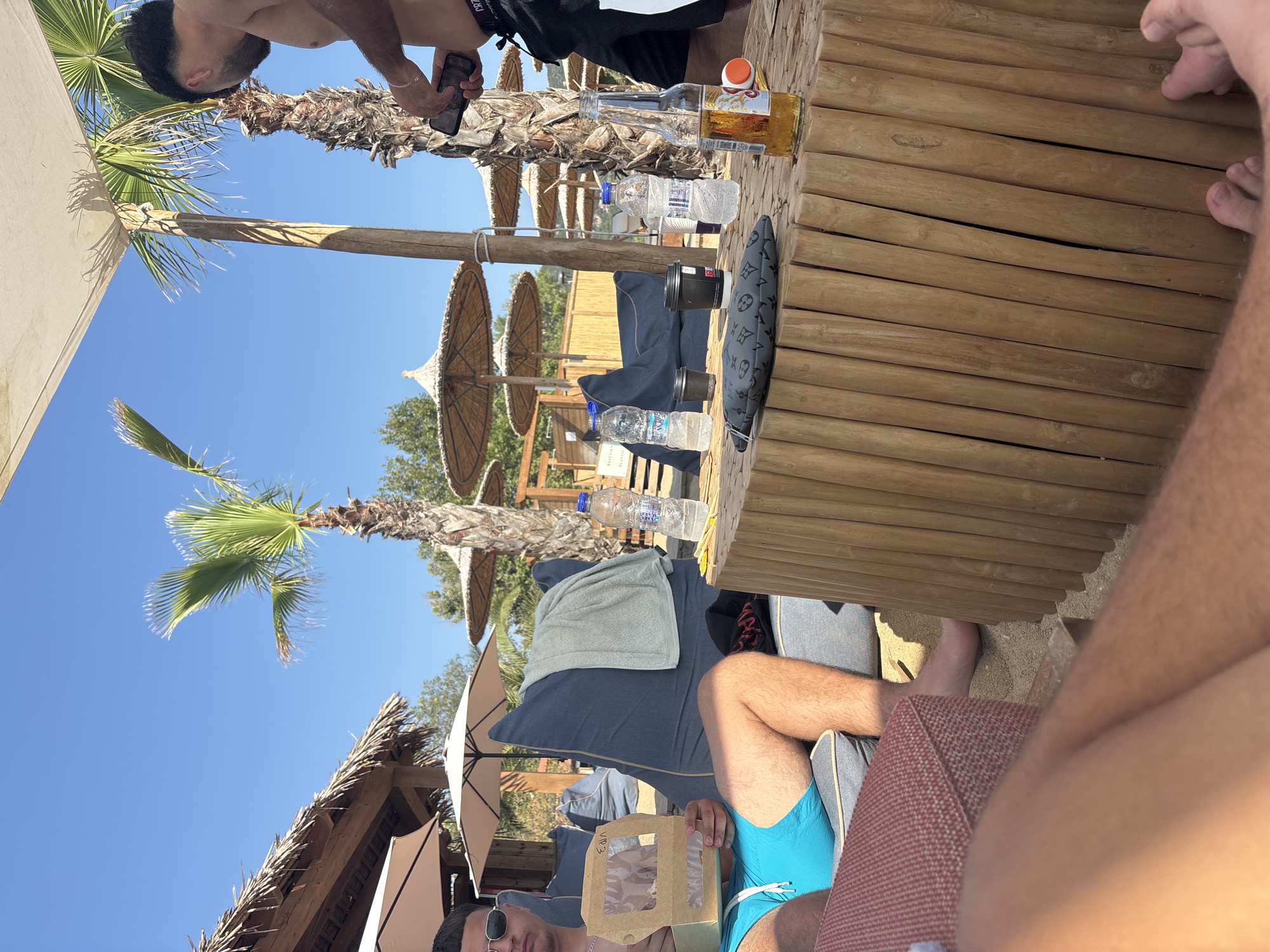 The image depicts a scene at a beachside establishment in Nea Irakleitsa, Greece. The setting appears to be an outdoor seating area, likely a beach bar or restaurant, characterized by sandy ground, palm trees, and thatched umbrellas providing shade. Two men are visible, one seated on a large, dark blue beanbag chair, wearing sunglasses and blue swimming trunks, and holding a small takeout container. He appears to be eating or preparing to eat from it. The second man, standing, is wearing black shorts and is partially visible as he holds a mobile phone. In the foreground, a wooden table is laden with several items, including two disposable coffee cups, two bottles of water, and a bottle of beer. A dark grey, patterned clutch or pouch rests on the table, alongside some small wooden skewers. The overall atmosphere suggests a relaxed day at the beach.