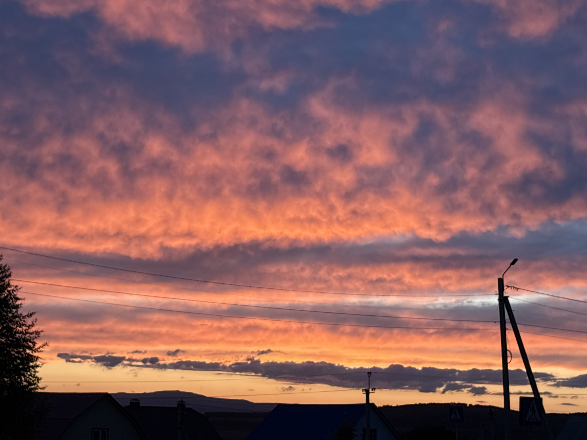 The image captures a dramatic sunset in an unidentified location, likely a town or residential area. The sky is filled with colorful clouds, with hues of orange, pink, and darker blue/gray shades. These clouds are layered and somewhat patterned, adding visual interest to the upper portion of the image.
In the foreground, there are rooftops of several houses, with varying colors and shapes. A few distant hills are also visible, adding a sense of depth to the landscape. A utility pole with power lines stretches across the scene, a common feature in residential areas. A tree is seen on the left edge of the frame. The light suggests that the sun is setting behind the houses, creating a silhouette effect for the buildings and the utility pole.