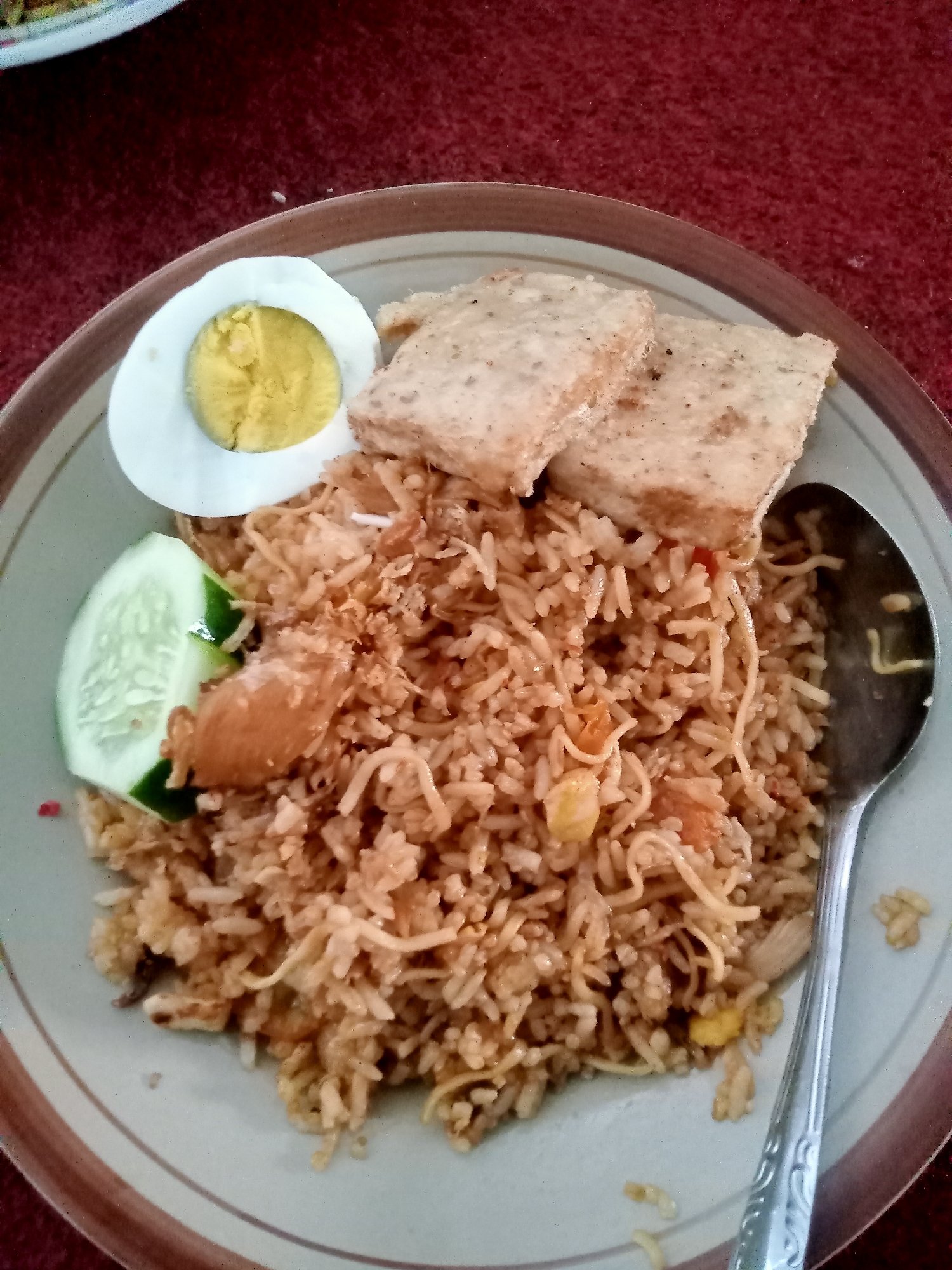 A close-up shot shows a plate of Indonesian fried rice, likely Nasi Goreng, served with accompaniments on a red tablecloth. The fried rice appears to be mixed with small pieces of meat, possibly chicken, and some vegetables. On the side, there is a slice of hard-boiled egg, two pieces of fried tofu, and a slice of cucumber. A silver spoon rests on the right side of the plate, ready for use. The plate has a brown rim. The background is a red textured surface, possibly a tablecloth or placemat. There is no visible indication of the specific location within the city of Simo, Indonesia.