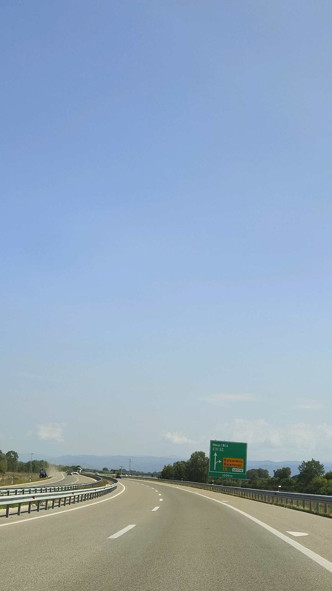 The image shows a highway leading through a rural landscape under a clear blue sky. On the right side of the road, a green directional sign indicates "Ниш / Niš" with an arrow pointing straight, and "Бујановац / Bujanovac" with route number 41 and an arrow pointing right. Below this, it indicates "ЦЕНТАР / CENTER" with a distance of "1000m". The highway has multiple lanes and is bordered by a metal guardrail. In the distance, mountains are visible, and there are scattered trees and vegetation along the roadside. A few vehicles are present on the highway, including a truck in the distance. The scene suggests a location in or near Bujanovac Municipality, Serbia, along a major road.