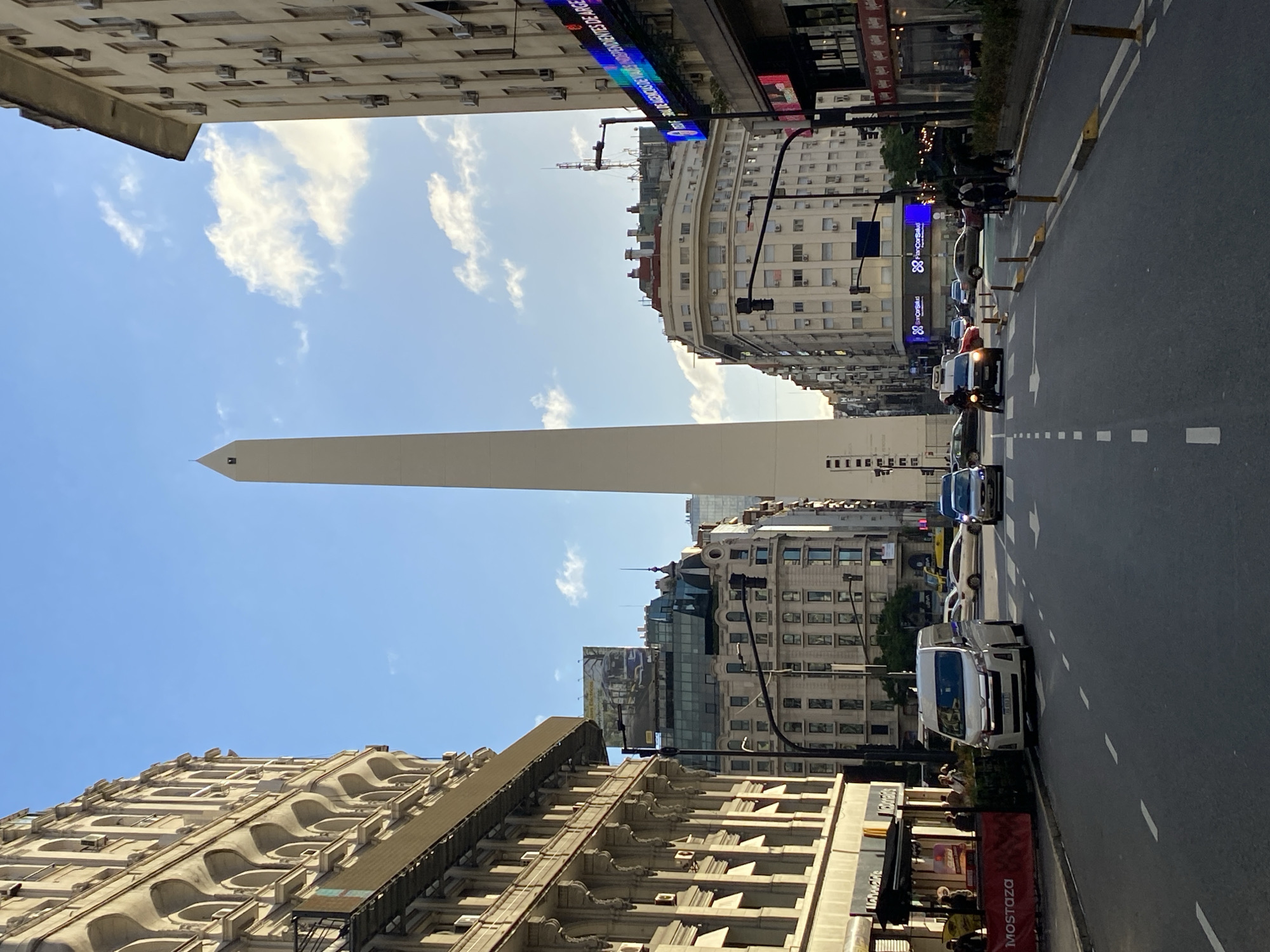 The image captures a vibrant street scene in Buenos Aires, Argentina, dominated by the iconic Obelisco de Buenos Aires, a tall, slender white monument, rising vertically towards a bright blue sky with scattered white clouds. The photograph is taken from a low angle, giving a sense of looking up from street level, and is oriented vertically, showcasing the height of the surrounding buildings.
On either side of the wide street, multi-story buildings of varying architectural styles line the thoroughfare. On the left, a grand, ornate building with decorative cornices and balconies stands, while on the right, structures include a more modern building with numerous windows and air conditioning units. A large, curved building is visible further down the street on the right.
The street itself is wide, with clear white lane markings and yellow bollards separating lanes or pedestrian areas. Several vehicles are visible, including cars and a white van, suggesting active traffic or parked cars. A few people can be seen on the sidewalks, adding to the urban feel.
Visible text on signs and digital displays provides further context. A blue LED screen on a building displays scrolling text, partially legible as "DE MERCADO DE CAPITALES DE INVERSORES DE ARGEN...", likely referring to financial market information. Another sign features the logo and name "Sancor Salud," indicating a health insurance provider. Closer to the foreground on the right, a red banner displays "Mostaza," identifying a local fast-food chain. The bright daylight suggests it's daytime, with clear weather conditions enhancing visibility of the city's architecture and the towering obelisk.