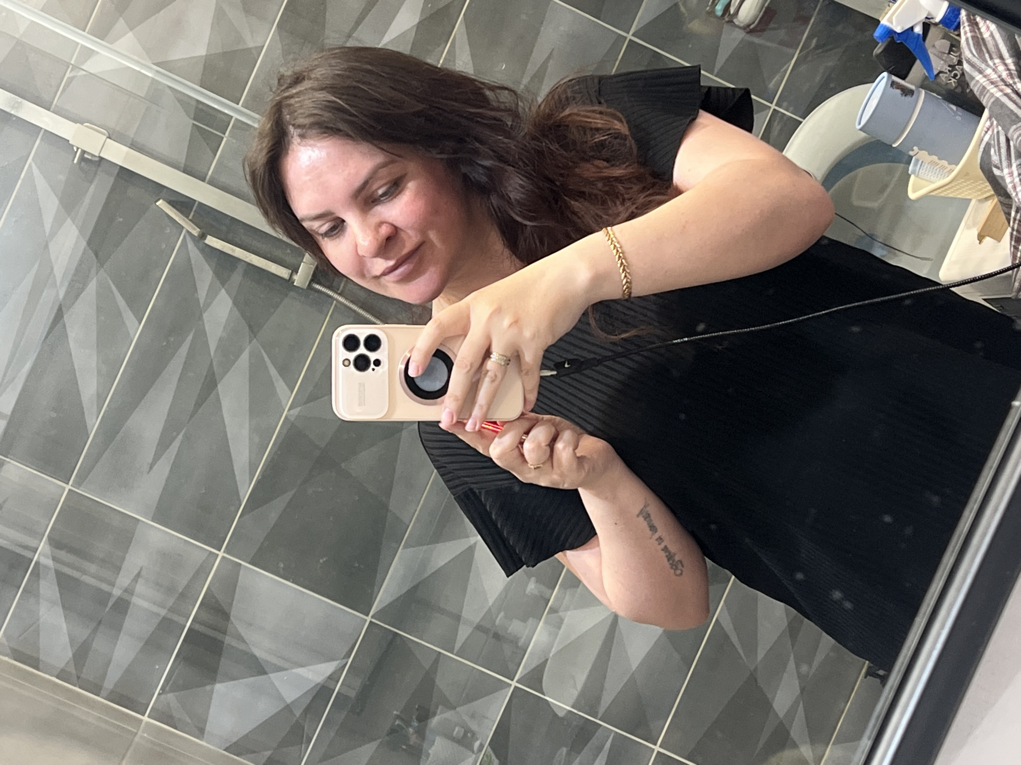 A woman is taking a selfie in a bathroom. She is holding a phone in her right hand, and the phone has a light-colored case with a dark circle on it. The woman is wearing a black dress and a gold bracelet on her left wrist. She has brown hair and fair skin. The bathroom has gray tiled walls and floors. In the background, there is a toilet with cleaning supplies on it. The location in the city of Noviy, Russia is undetermined.