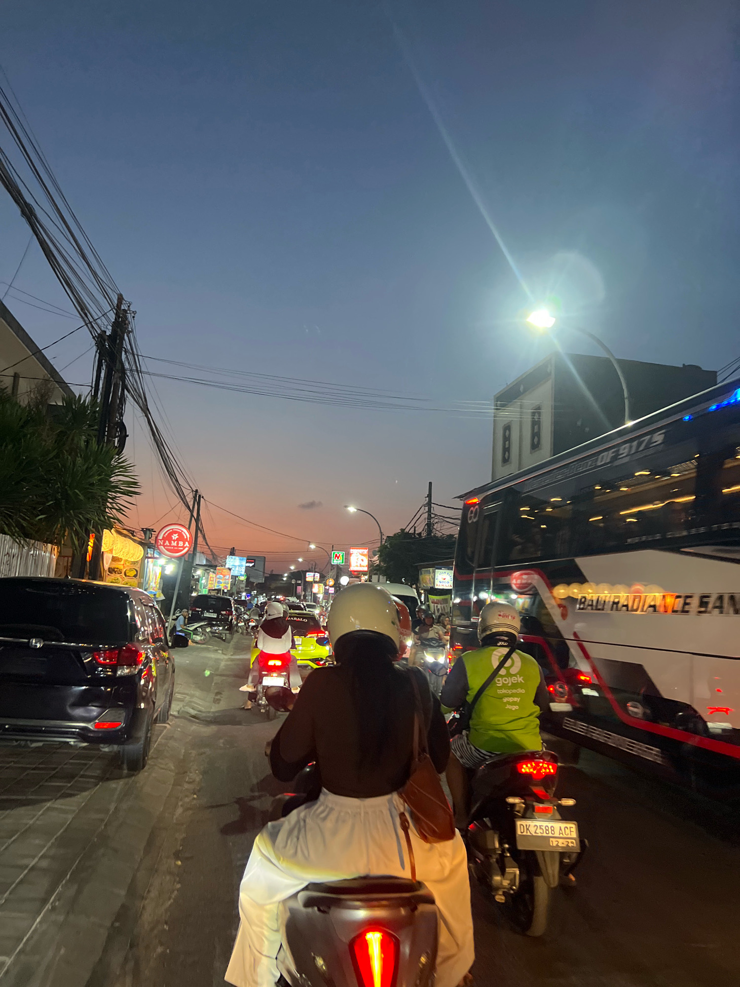 This is a full, eye-level, vertical shot taken in Kuta, Indonesia, likely during the evening. The sky is a gradient of deep blue fading into orange and pink hues near the horizon, indicative of sunset or twilight. The scene depicts a busy street with heavy traffic, primarily motorcycles and cars.
In the foreground, a person on a motorcycle is seen from behind, wearing white pants and a dark top. They are followed closely by another motorcycle, also seen from behind, with a rider wearing a yellow helmet and a green Gojek-branded vest over striped shorts. This motorcycle has a license plate reading "DK 2588 ACF".
To the left of the foreground motorcycle, a black SUV is stopped in traffic. Further down the street, other vehicles, including a bright yellow car and more motorcycles, are visible. Several shops and businesses line the street, with illuminated signs and storefronts. One sign prominently displays "NAMBA".
Dominating the right side of the frame is a large bus with the text "BALI RADIANCE SAN" visible on its side. The bus has multiple illuminated windows, and the overall impression is that of a congested urban environment during peak traffic hours. Overhead, a complex network of electrical wires stretches across the sky, crisscrossing above the street. A bright street lamp illuminates a section of the sky to the right, creating a lens flare effect. The road surface appears to be asphalt with some visible debris or unevenness.
