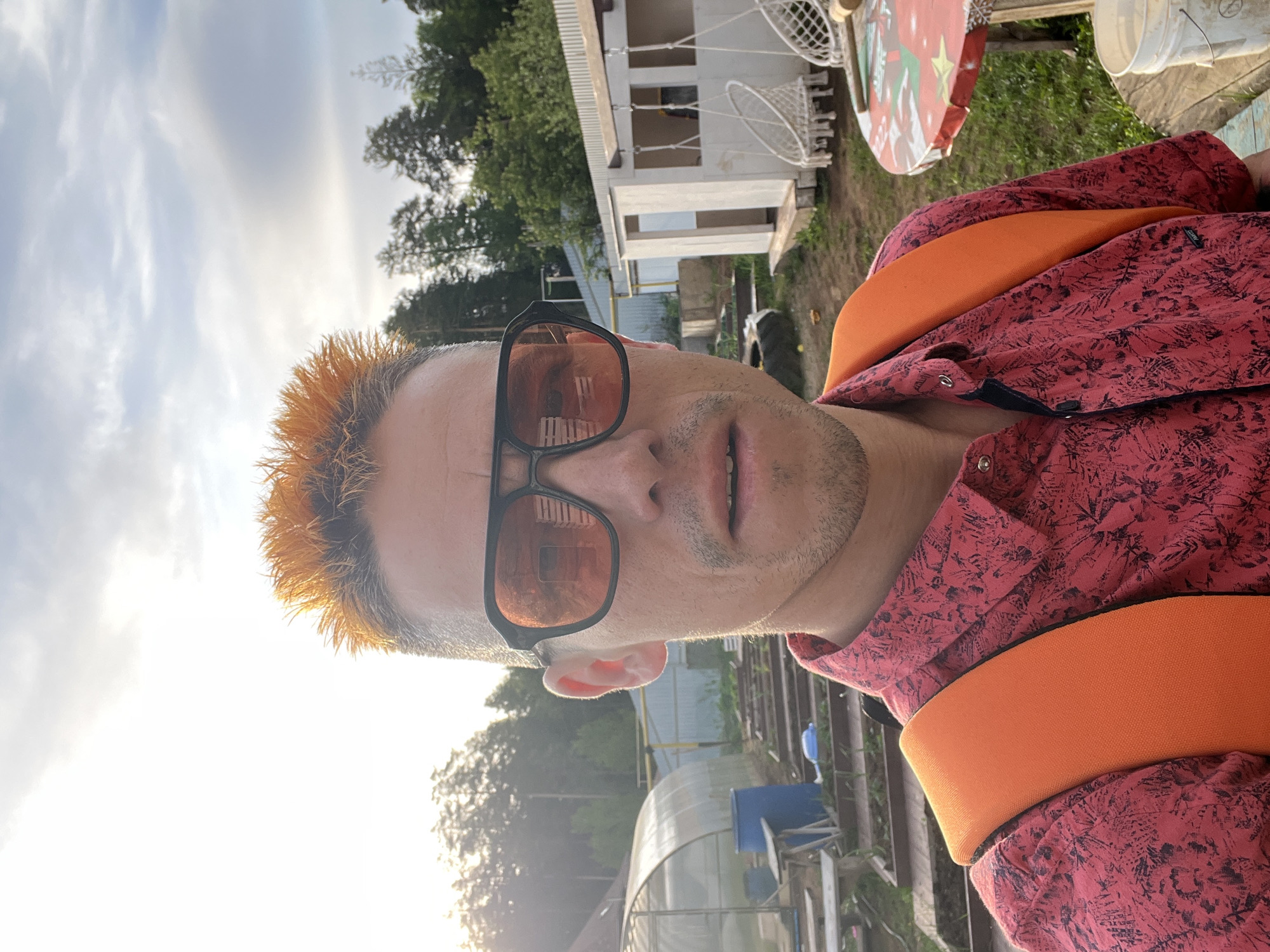 A selfie of a man with spiked, orange-colored hair wearing sunglasses and a red patterned shirt with orange straps. The background shows a house, trees, a greenhouse, and the sky with sunlight. Given the location of the person involved, this could be a suburban or rural area in the city of Chaikovsky, Russia.