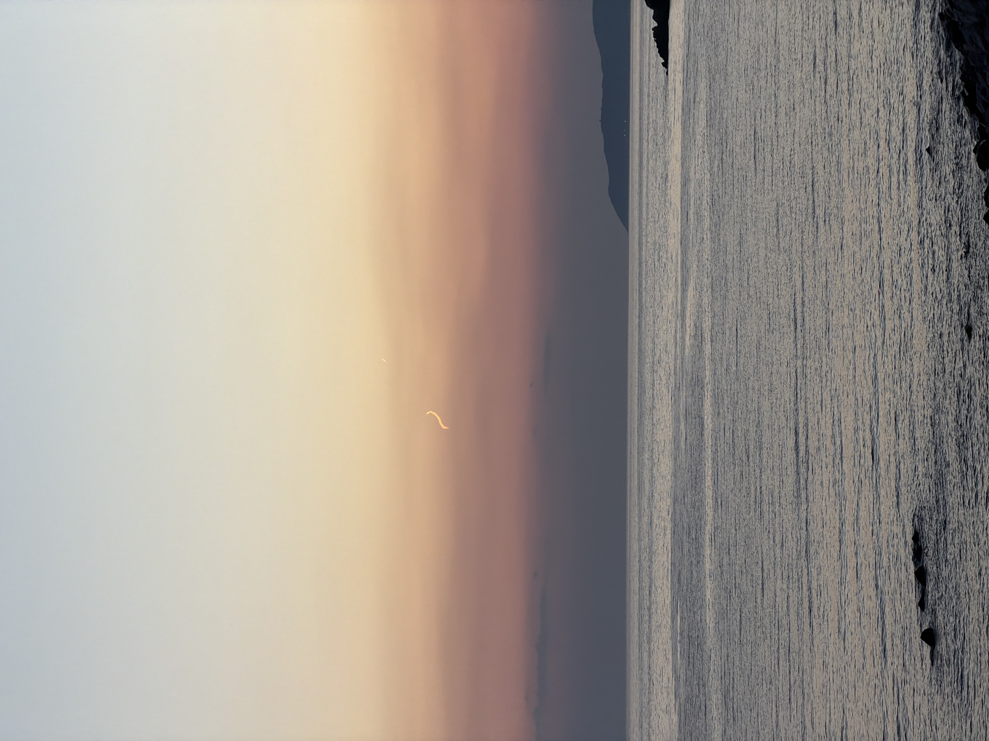 The image depicts a serene seascape at dusk in Perazića Do, Montenegro. The sky exhibits a soft gradient of colors, transitioning from pale blue at the top to warm orange and pink hues closer to the horizon. A faint, wispy cloud formation is visible in the mid-sky, with a delicate, curved streak of light, possibly from an aircraft or a celestial phenomenon, also present.
The sea stretches out before the viewer, appearing calm with subtle ripples and reflections of the sky's colors. In the foreground, a rocky shoreline is visible, with dark, weathered stones partially submerged in the water. In the distance, the silhouette of a landmass, likely a hill or mountain, is discernible against the twilight sky, with a few faint lights possibly indicating a settlement. The overall impression is one of tranquility and natural beauty, characteristic of the coastal landscape of Perazića Do. There are no people or animals visible in the scene.