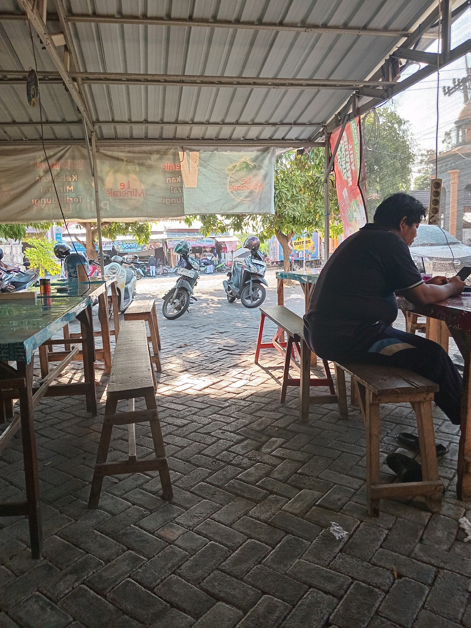 A man sits at a wooden table on a paved outdoor patio in Kembangan, Indonesia. He is wearing a dark t-shirt and dark pants with blue stripes. He is looking at his phone, which he holds in his right hand. A plastic cup with a dark liquid is on the table in front of him, along with a crumpled white napkin. The patio is covered by a corrugated metal roof supported by wooden beams and poles. There are several wooden tables and benches on the patio. In the background, motorcycles are parked. Beyond the patio, there are trees and buildings. The overall scene suggests a casual dining or rest stop area.