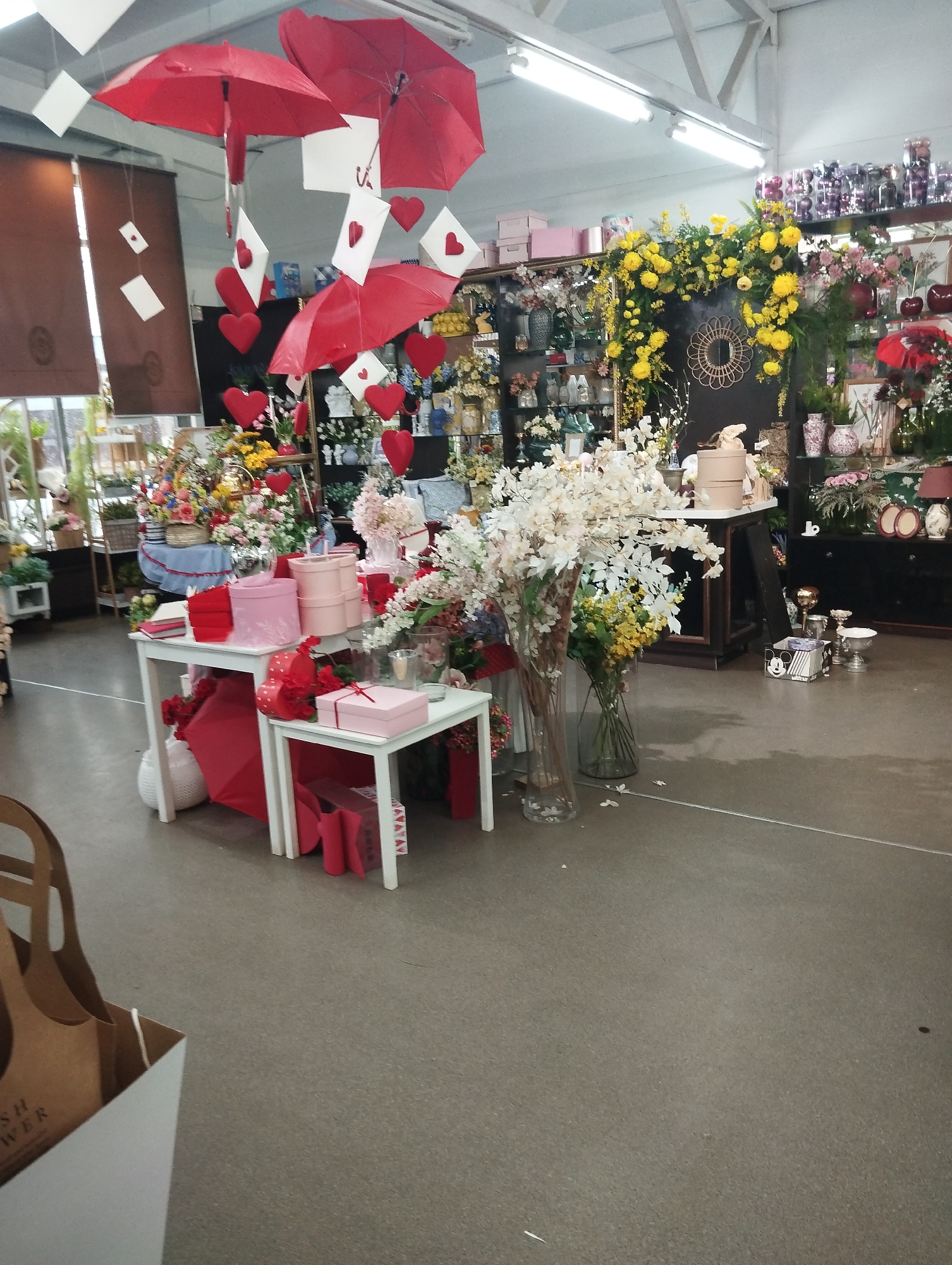 The image shows the interior of a shop, likely a flower or gift shop, possibly in Votkinsk, Russia. Valentine's Day decorations are prominently displayed, including red umbrellas suspended from the ceiling with attached hearts and envelopes. Two white tables display pink gift boxes, red ribbons, and additional decorations. Vases of flowers, including white blossoming branches, are placed on the floor. In the background are shelves stocked with various vases, flowers, and other decorative items. A decorative archway of yellow flowers frames a section of the shop. The floor is gray and smooth. The lighting is bright, likely from overhead fluorescent fixtures. A partial view of a shopping bag is visible in the foreground.