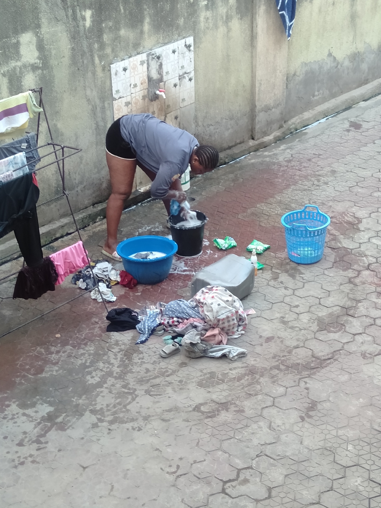 An outdoor scene in Shangisha, Nigeria, shows a woman engaged in handwashing laundry. She is positioned centrally, bent over, with her hands immersed in a black bucket filled with soapy water and clothes. To her right, a blue plastic basin also contains water and clothes, likely for rinsing.
The woman has dark skin and braided hair, and is dressed in a grey long-sleeved shirt and black shorts with a white trim. She is wearing light-colored slip-on sandals.
The ground is paved with hexagonal interlocking stones and is visibly wet, indicating active washing. To the left, a metal clothes drying rack holds some garments, including a striped towel and dark clothing. Various other clothes are scattered on the wet pavement around the washing area.
In the background, a textured concrete wall features a section of white patterned tiles with a red faucet. Several items are on the ground near the woman: a grey plastic jerrycan, two small green plastic sachets (possibly detergent), a white bottle with a pump dispenser, and an empty blue plastic laundry basket further to the right. Some footwear, including sandals, is mixed with the piles of clothes on the ground.
The scene appears to be taking place during the daytime, under soft lighting, possibly indicating an overcast sky or early morning/late afternoon. No discernible text is visible in the image.