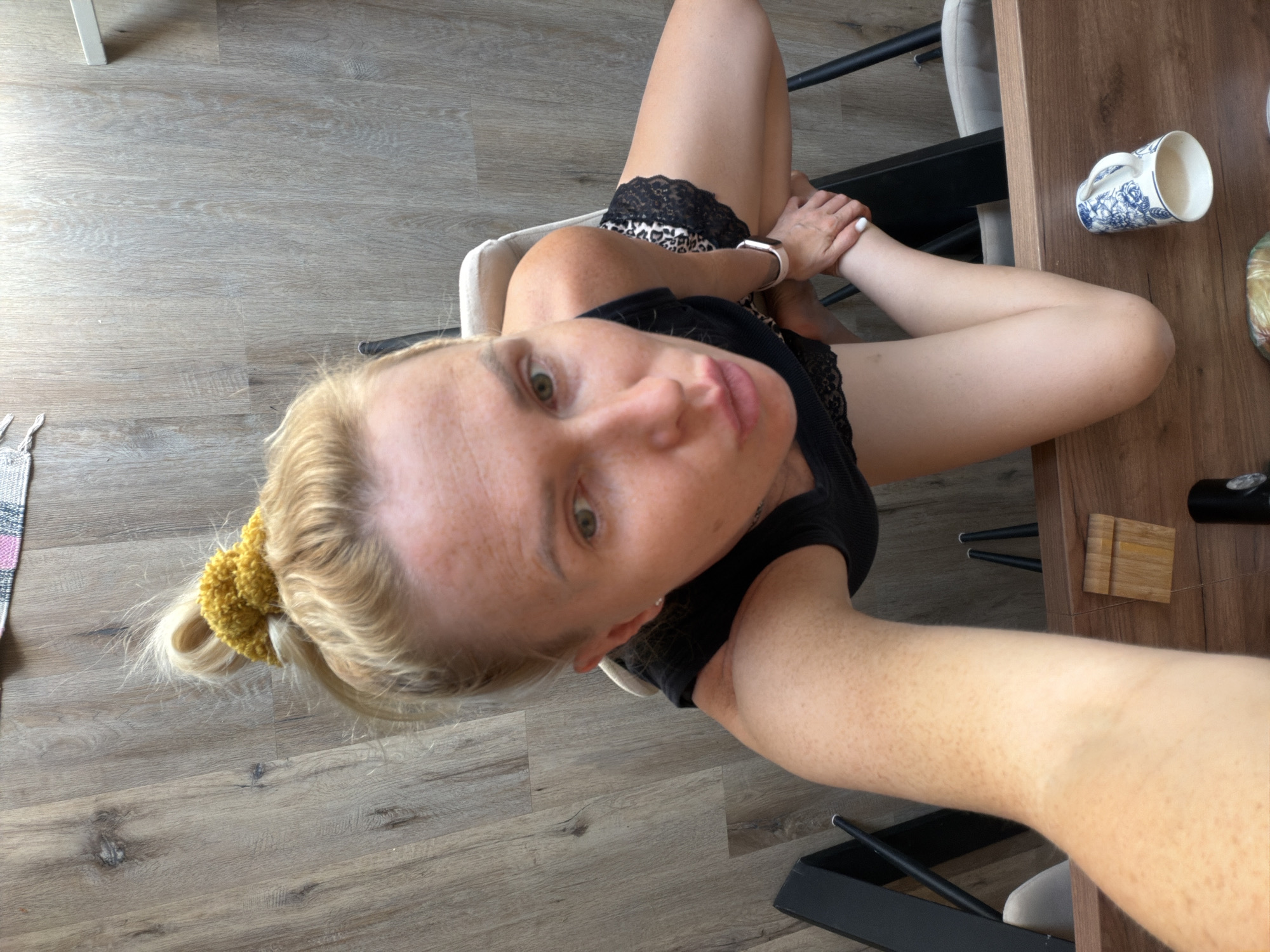 A woman is seated at a wooden table, her legs crossed, wearing a black lace-trimmed top and leopard-print shorts. She is holding her hand to her leg, and a mug with a blue floral pattern sits on the table beside her. The scene is indoors, with a wooden floor and a portion of a dark-colored chair visible. The perspective is from above and slightly to the side, capturing the woman's face and upper body.