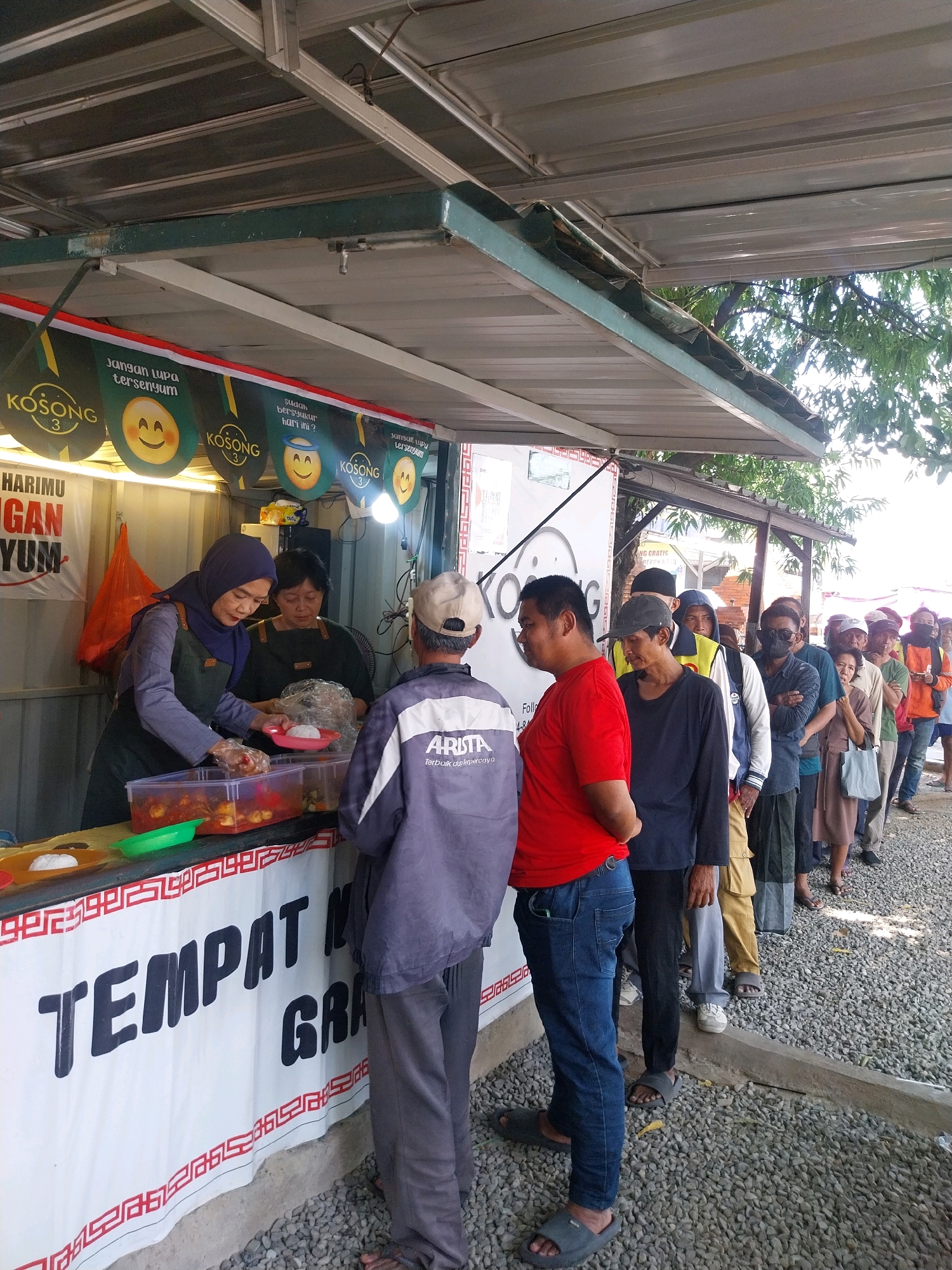 This is a street-level, eye-level, medium shot photograph taken outdoors in Cirebon, Indonesia. The scene depicts a food stall with a queue of people waiting to be served. The food stall is set up under a metal awning, with a banner behind the counter that reads "TEMPAT NONGKRONG GRATIS" (Free Hangout Spot). Several smaller banners with smiling emoji faces and the word "KOSONG" are also visible, suggesting a brand or promotion.
Two women are working behind the counter. One woman, wearing a purple hijab and a green apron, is scooping food from a large plastic container into a pink plate. The other woman, wearing a dark green apron, is standing next to her, holding a plastic bag. In front of the stall, a man in a grey jacket and a man in a red t-shirt are interacting, possibly discussing their order or waiting their turn.
A line of approximately ten to fifteen people, mostly men, are waiting patiently. They are dressed in casual clothing, and some are wearing hats or masks. The ground is covered in gravel. The overall impression is that of a busy, informal food service operation catering to the local community.