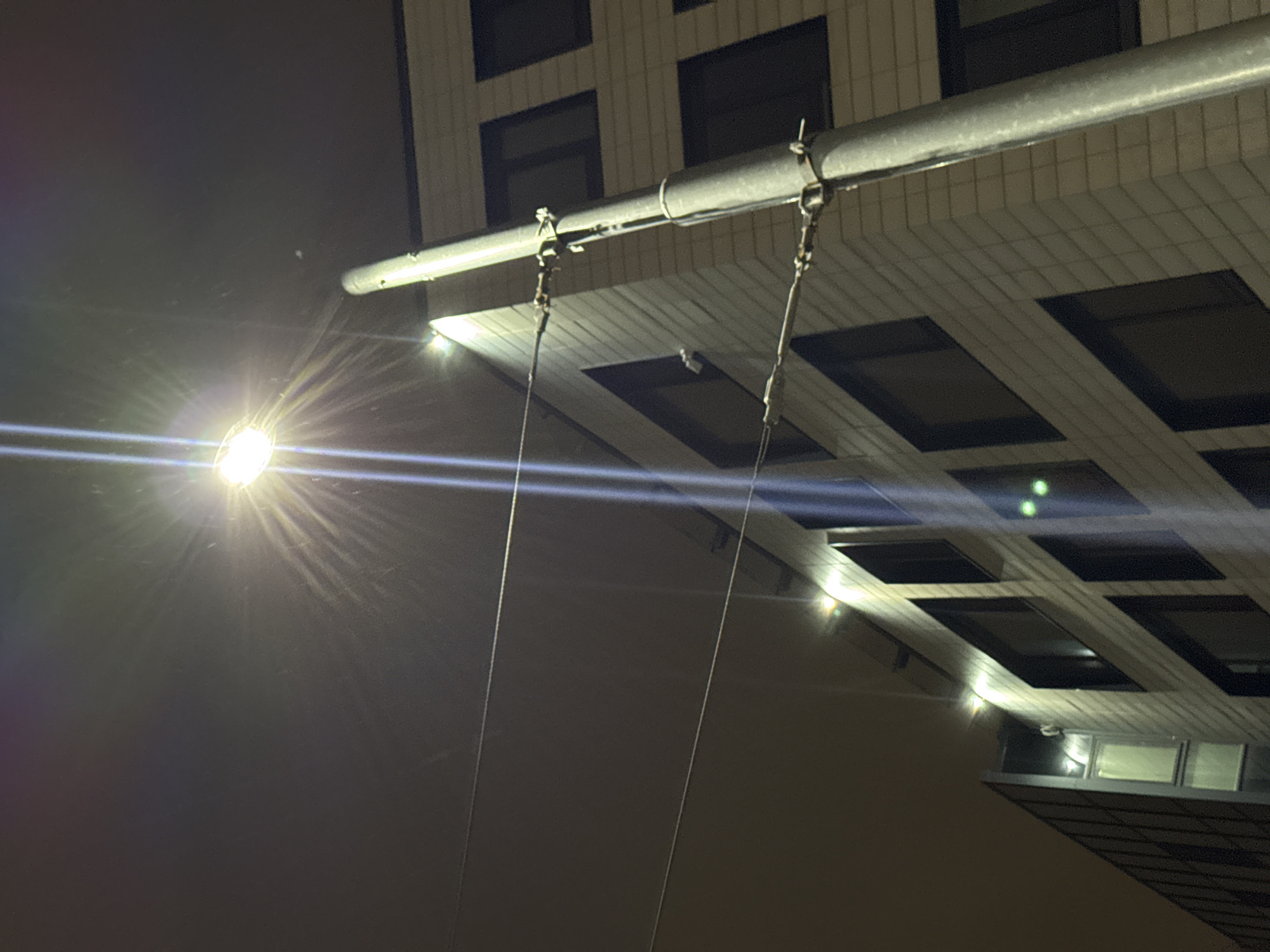 Captured at night, the image presents a low-angle view of a building facade in what appears to be Sofia, Bulgaria. A metal pipe runs horizontally across the upper part of the frame, anchored to the building's overhang by wires. The overhang features a grid pattern of recessed dark squares, likely windows or architectural features. A bright light source emanates from the left side, casting beams across the scene and creating a strong lens flare effect. The surrounding environment appears dark.