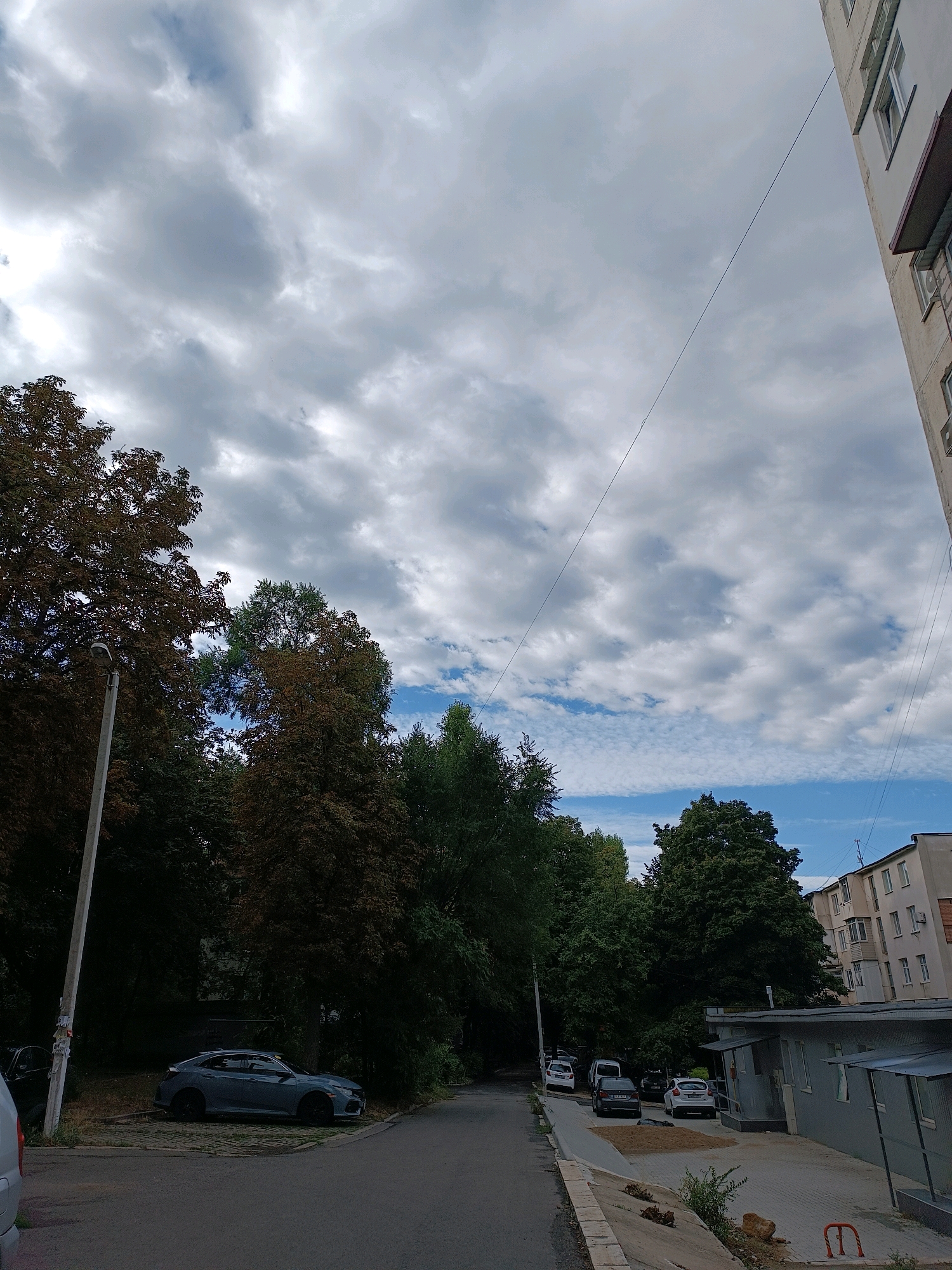 A residential street scene in Chișinău, Moldova, captured on a cloudy day. The view is angled upwards, with a large expanse of sky filled with dense, grey clouds and patches of blue. On the right side of the frame, the corner of a light-colored, multi-story apartment building is visible, featuring rows of windows and some architectural details. A thin wire extends diagonally across the sky from the building.
On the left side of the street, a tall, grey lamppost stands near a sidewalk. Behind the lamppost, a cluster of trees with dark foliage forms a dense canopy. A light blue car is parked on the side of the road in front of the trees. The street itself is paved and appears to be asphalt.
Further down the street, more trees are visible, with a mix of dark green and reddish-brown foliage. Several parked cars are scattered along the sidewalk. A low, grey, single-story building with a covered entrance and a paved area in front of it is situated on the right side of the road, behind the apartment building. The ground in front of this building is covered with interlocking pavers. There is a pile of dirt and some debris on the paved area. A red metal object, possibly a bollard or part of a bike rack, is visible in the lower right corner. The overall impression is that of a somewhat overcast, urban environment with interspersed greenery.