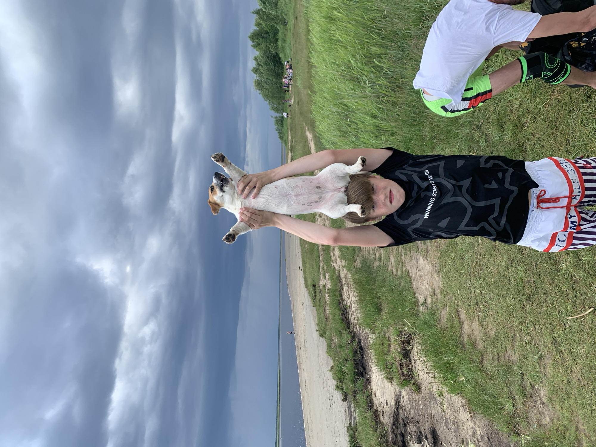 A young boy stands in a grassy area near a beach, holding a Jack Russell Terrier dog above his head. The dog is upside down, with its paws in the air. The boy is wearing a black t-shirt and white shorts with red and blue stripes. Another person, partially visible, is wearing a white t-shirt and neon green shorts, with a black knee brace. In the background, there is a beach, a body of water, and a line of trees. The sky is overcast with dark clouds. The image was taken in Severodvinsk, Russia.
