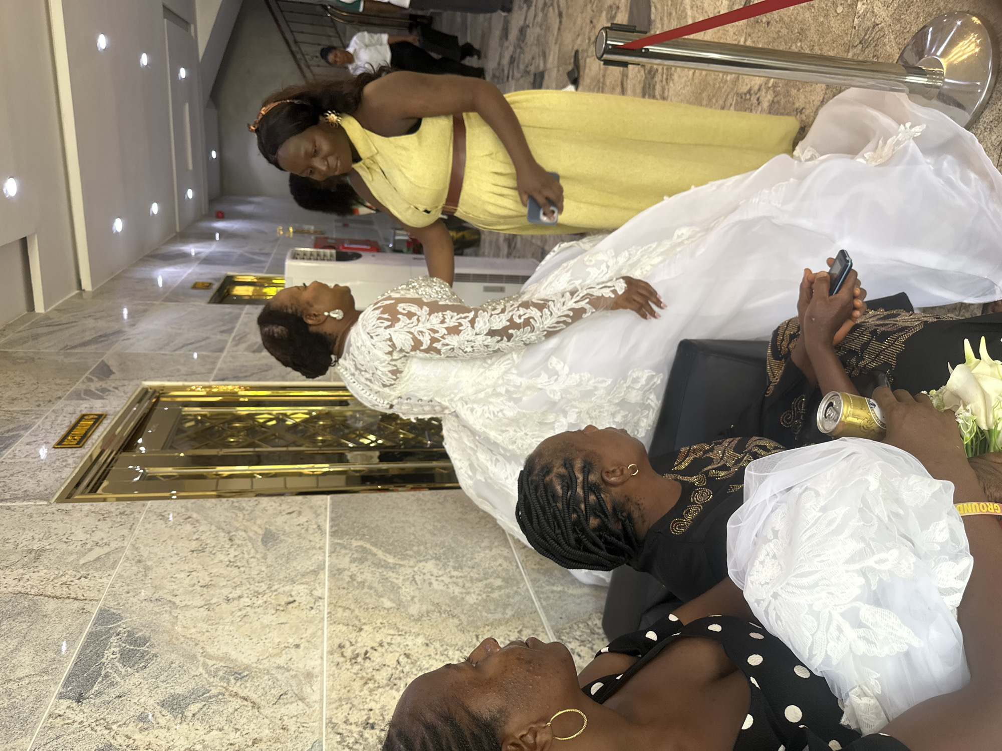 In a well-lit indoor setting, possibly a reception hall or event space, three Black women are the primary subjects. One woman, adorned in a white lace wedding dress, is seated and looking upwards. Another woman, wearing a yellow dress and a headband, is standing and looking down at the bride, holding a cell phone. A third woman, in a black dress with gold embellishments, is also seated and looking upwards, holding a can and a bouquet of white flowers. The floor is tiled with a marble-like pattern, and there is a golden decorative doorway or panel in the background. A red rope barrier is visible, suggesting a designated area or queue. The overall atmosphere appears to be festive and celebratory, likely related to a wedding ceremony or reception. The lighting suggests it is daytime, and there are no visible weather cues. The location context is Abuja, Nigeria. A yellow sign with black text that reads "GROUND" is visible on the arm of the woman with the flowers.
