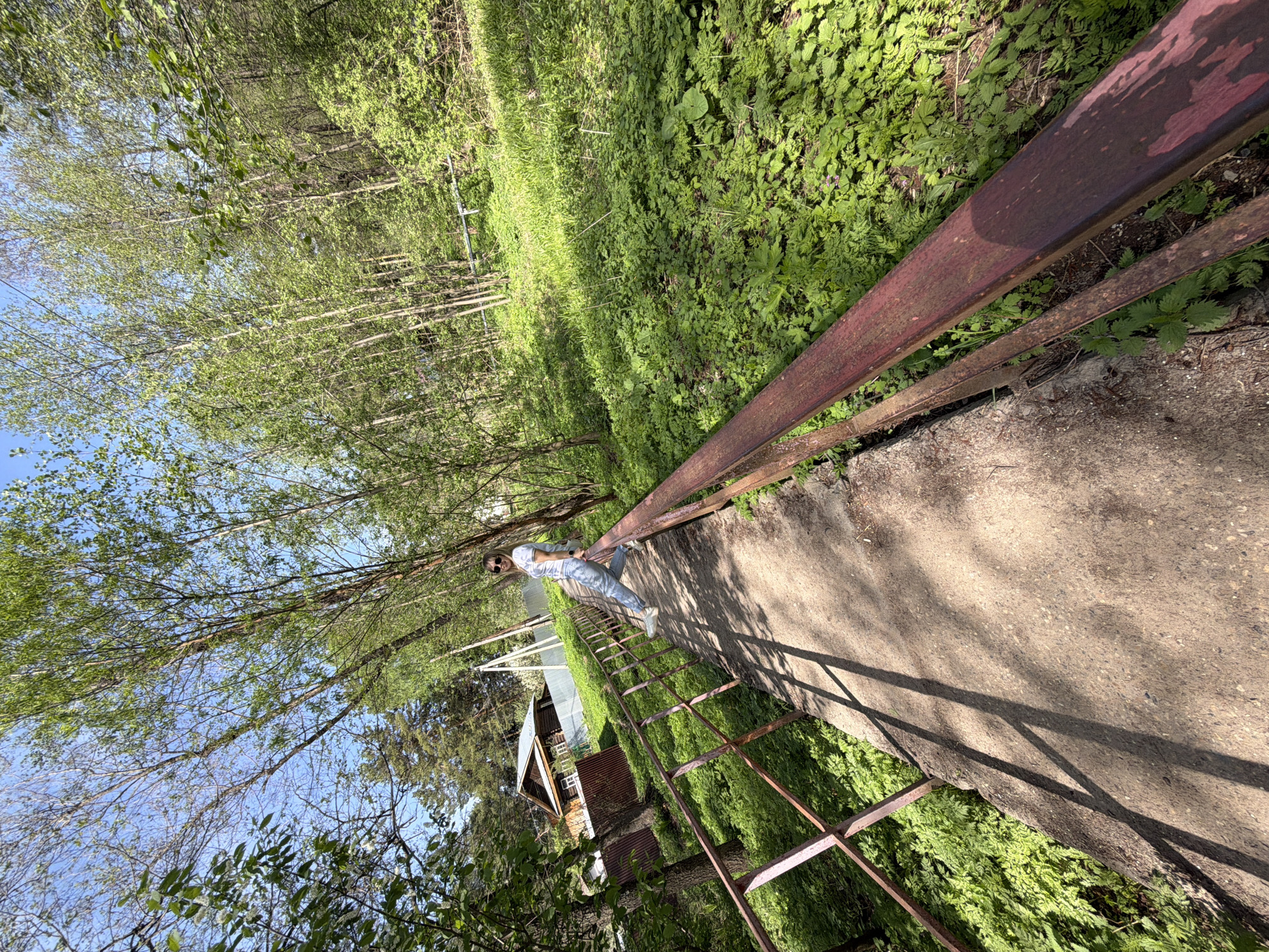 The image shows a person standing on a concrete walkway with a rusted metal railing. The walkway is flanked by dense green vegetation. A rusty metal ladder is present on the side of the walkway. Beyond the vegetation and trees, a small building is visible. The sky is blue with scattered trees. The setting appears to be outdoors in a natural, possibly wooded, environment. The location may be Chaikovsky, Russia.
