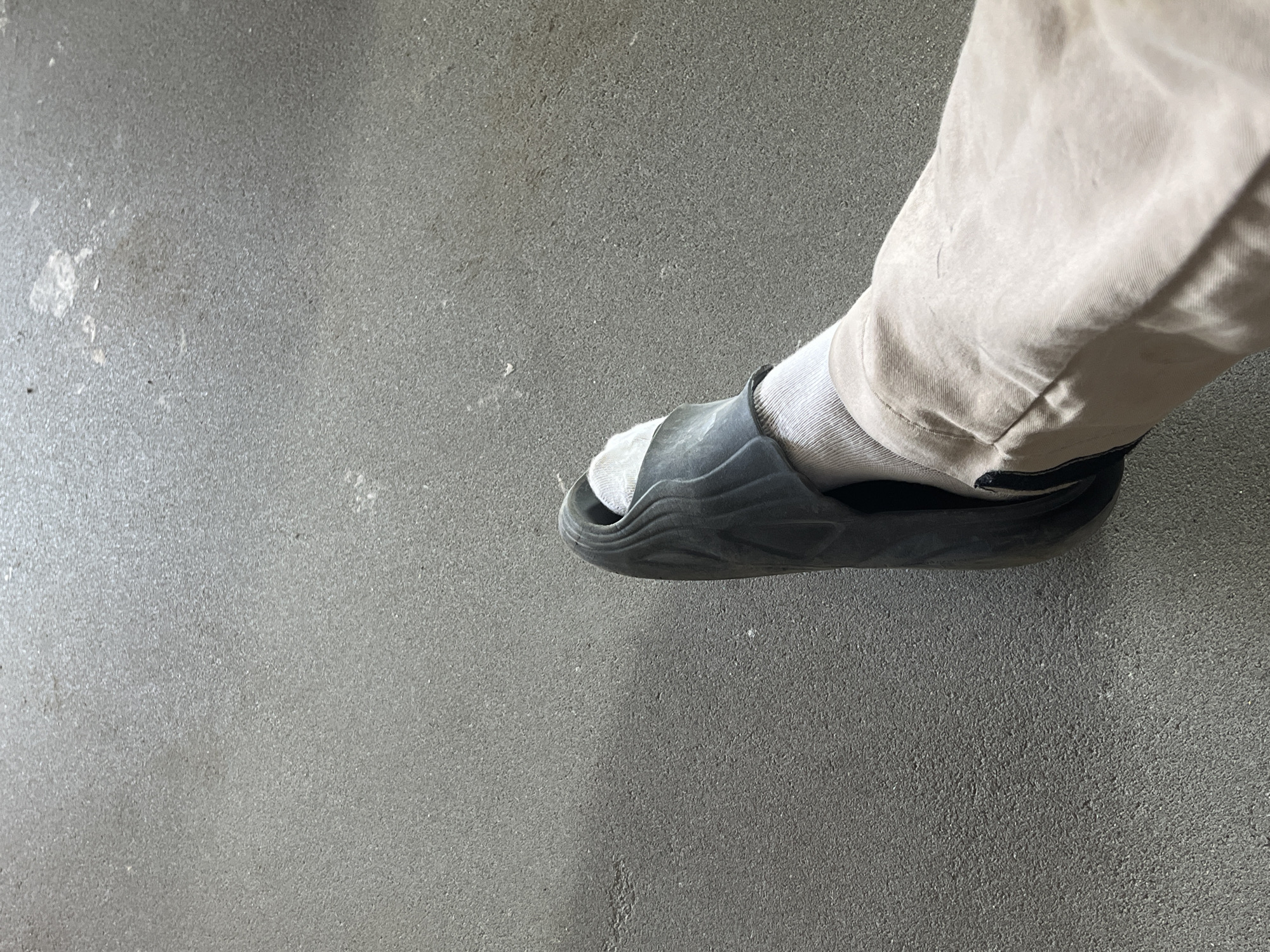 The image shows a close-up, overhead view of a person's left leg, clad in a light-colored, loose-fitting pant and a white sock. The leg is encased in a dark gray slide sandal. The scene appears to be outdoors, on a textured, dark gray surface, possibly asphalt or concrete. The lighting suggests it is daytime. The location is Strumivka, Ukraine.