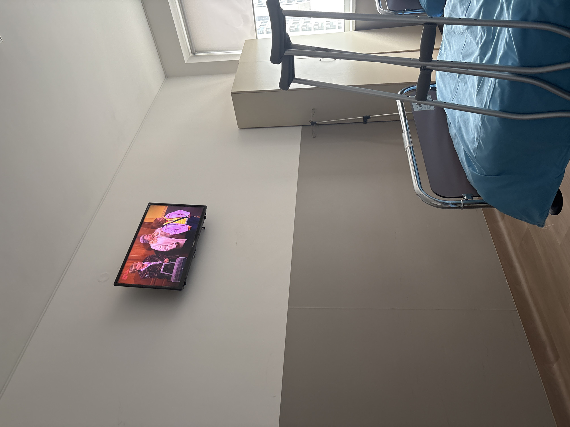 The image shows an interior space, possibly a hospital room in Sofia, Bulgaria. A television is mounted on a pale wall, displaying a show with several people on screen. To the right is a hospital bed, with a blue sheet. Overhead, natural light enters through a window. Crutches are positioned near the bed. The floor is a light wood tone. The wall is divided into two painted sections, the left wall is light and the right is gray.