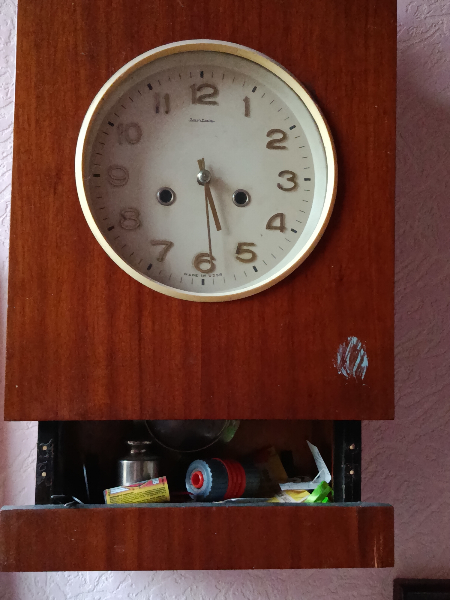 A wooden mantel clock with a white face and gold-colored trim is mounted on a textured pink wall. The clock features black Arabic numerals for hours and black tick marks for minutes. The hands are thin and metallic, with the hour hand pointing approximately to the 11 and the minute hand pointing to the 5. The brand name "Janus" is visible above the center, and "MADE IN USSR" is printed below the 6. Below the clock, there is an open compartment containing various items, including a cylindrical metal object, a yellow and red tube, a black and red cylindrical object, and some paper scraps. The compartment appears to be a drawer or shelf. The overall scene suggests a domestic setting, possibly a home in Смілавіцкі сельскі Савет, Belarus.