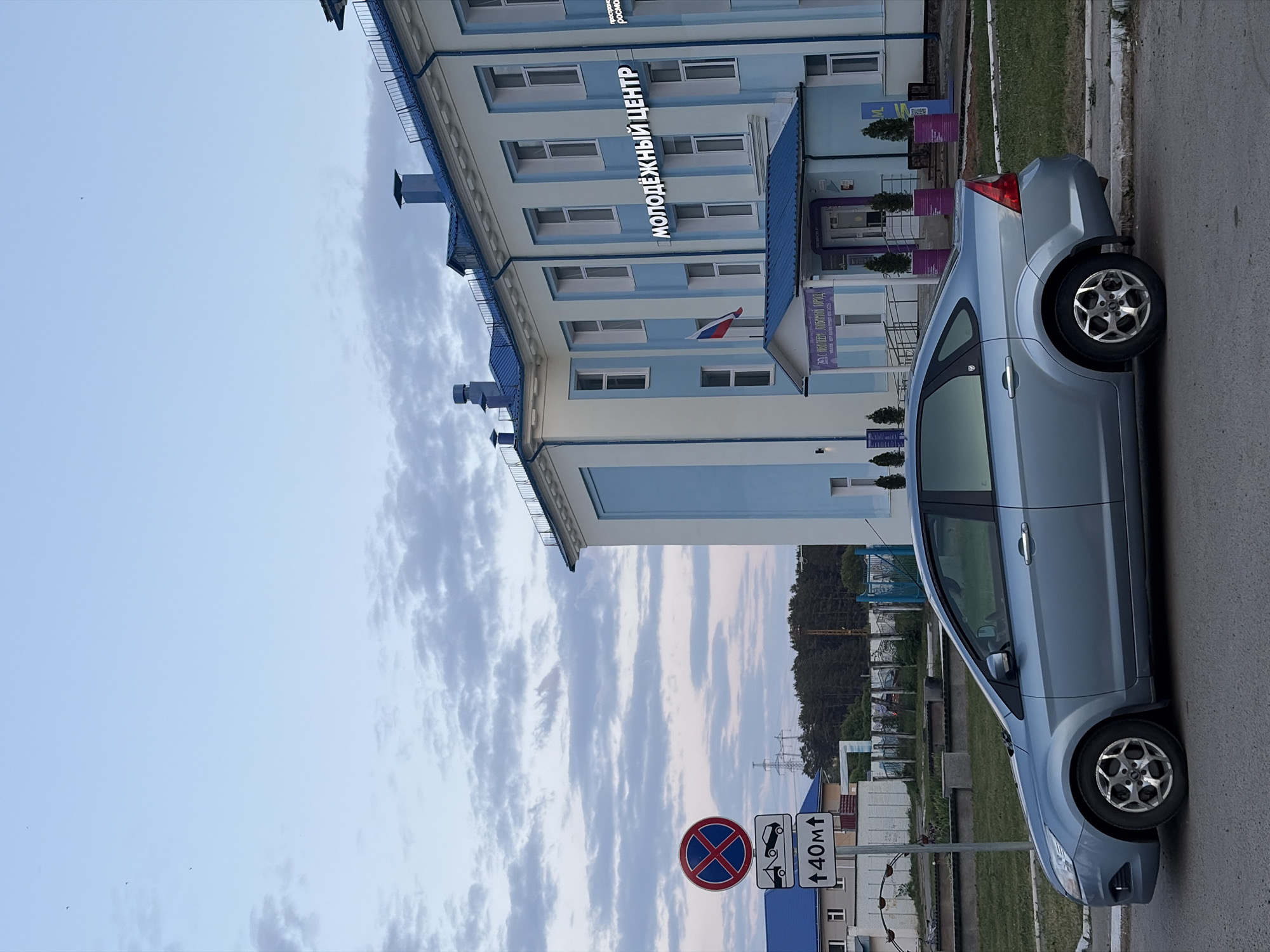 The image shows a light blue car parked on the right side of a street in Chaikovsky, Russia. The car is positioned near a building identified by a sign reading "МОЛОДЁЖНЫЙ ЦЕНТР" (Youth Center) in Russian. A Russian flag is visible on the building. A road sign indicates a no-stopping zone. Overhead power lines and trees can be seen in the background. The sky is visible with scattered clouds.