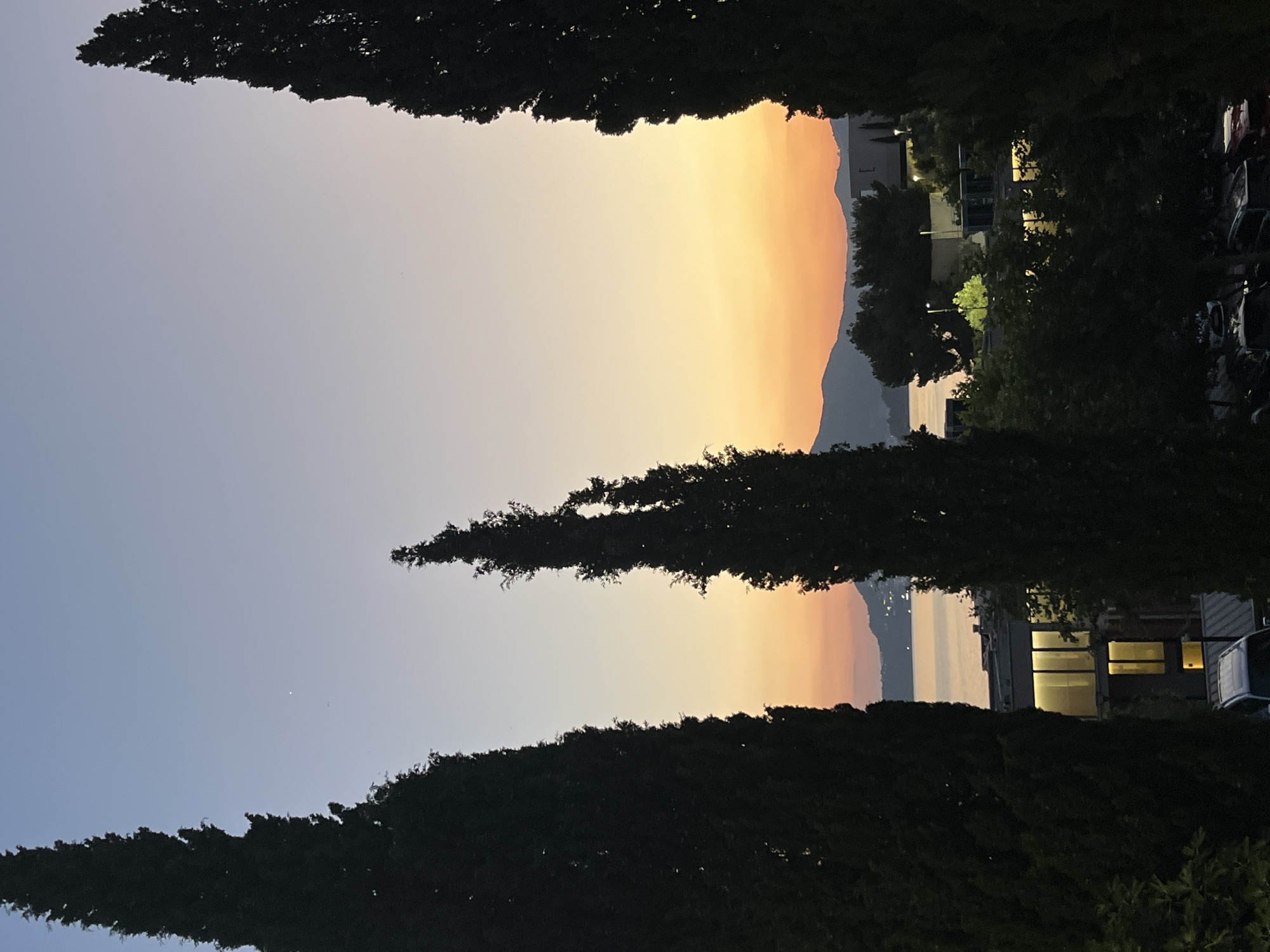 The image depicts a twilight scene in Pržno, Montenegro. Three tall, dark green cypress trees frame the view. In the background, a body of water is visible, with a silhouetted mountain range in the distance. The sky transitions from a pale blue at the top to soft shades of orange and pink near the horizon, indicating sunset or sunrise. Buildings and streetlights are visible in the mid-ground, suggesting an inhabited area. Several vehicles are parked in the foreground. The overall impression is one of a serene, natural landscape juxtaposed with human habitation.
