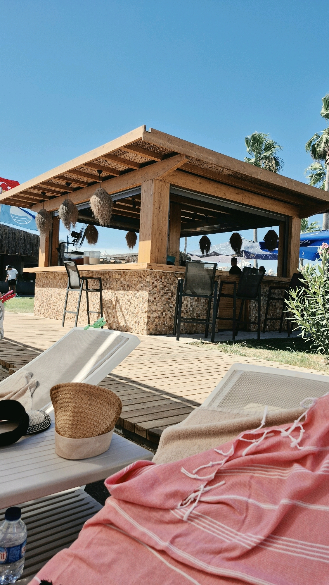 A beach bar with a wooden roof and stone facade is set against a clear blue sky in Serik, Turkey. The bar has a counter with several stools and is surrounded by a wooden deck. In the foreground, two empty lounge chairs are covered with towels. A straw hat and a bottle of water are placed on one of the chairs. Palm trees and lush green vegetation are visible in the background.