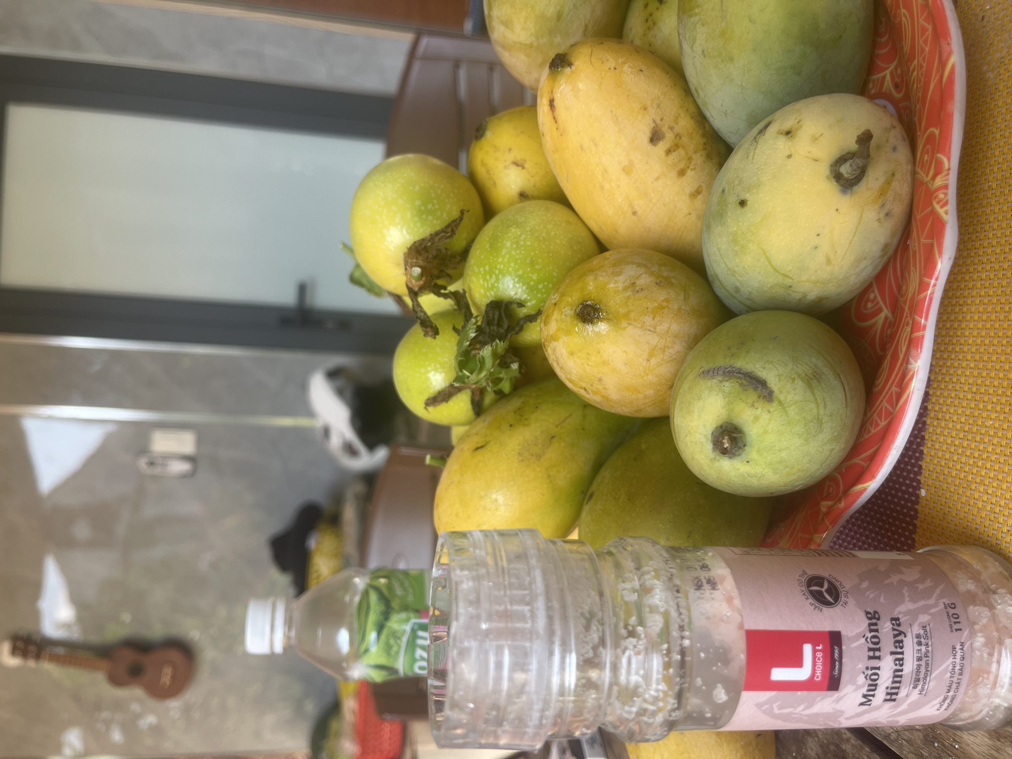 The image depicts a still life arrangement of fruits and a salt grinder on a wooden surface, likely outdoors in Phan Thiet Ward, Vietnam. A clear plastic bottle with a white cap, containing a green liquid and labeled "OZU," is positioned behind the salt grinder. The salt grinder, made of clear plastic, is filled with pink salt and has a label that reads "Muối Hồng Himalaya" in Vietnamese and Korean, indicating it is Himalayan Pink Salt. A vibrant red and white patterned plate holds a pile of ripe mangoes, varying in color from yellow to green, and several passion fruits, which are green with white speckles. In the background, a dark door frame with a closed door is visible, along with a wall and a decorative wooden ukulele hanging on it. The overall scene suggests a casual, domestic setting.