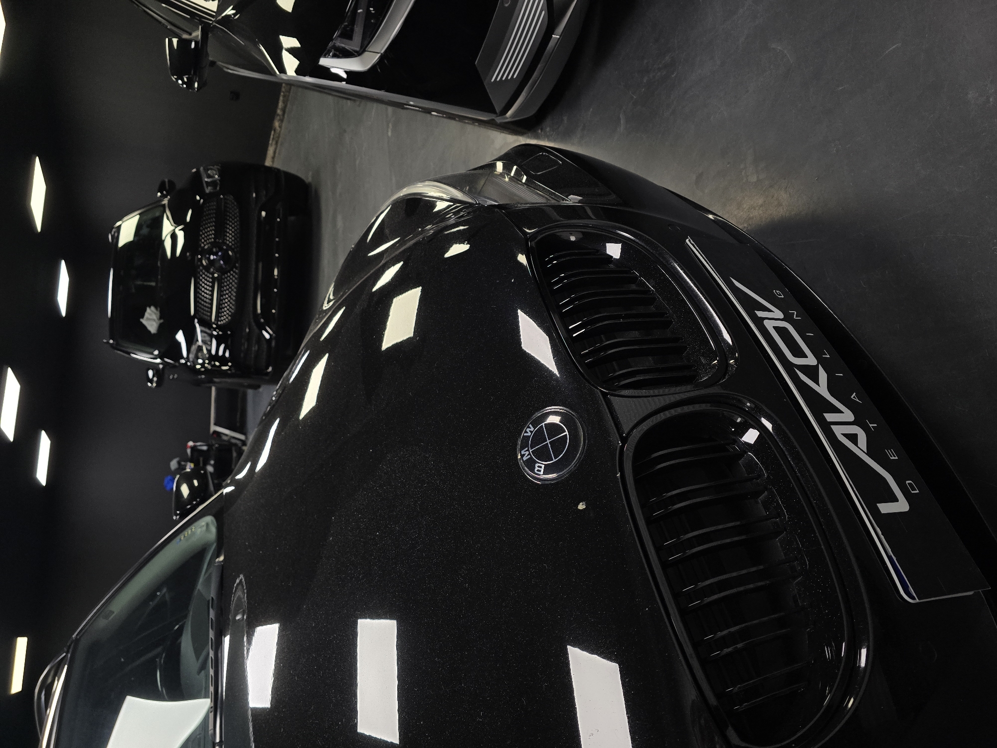 The image shows the front of a black BMW car, with its grille and headlights prominently featured. The car appears to be in a showroom or detailing studio, as evidenced by the polished floor and the bright overhead lighting. In the background, other black cars are visible, including a Mercedes-Benz SUV and another vehicle whose front is partially obscured. The scene is dark and modern, with a focus on the sleek lines and reflective surfaces of the cars. The overall impression is one of luxury and automotive care. There are no people visible in the image. The location is identified as Ruse, Bulgaria.