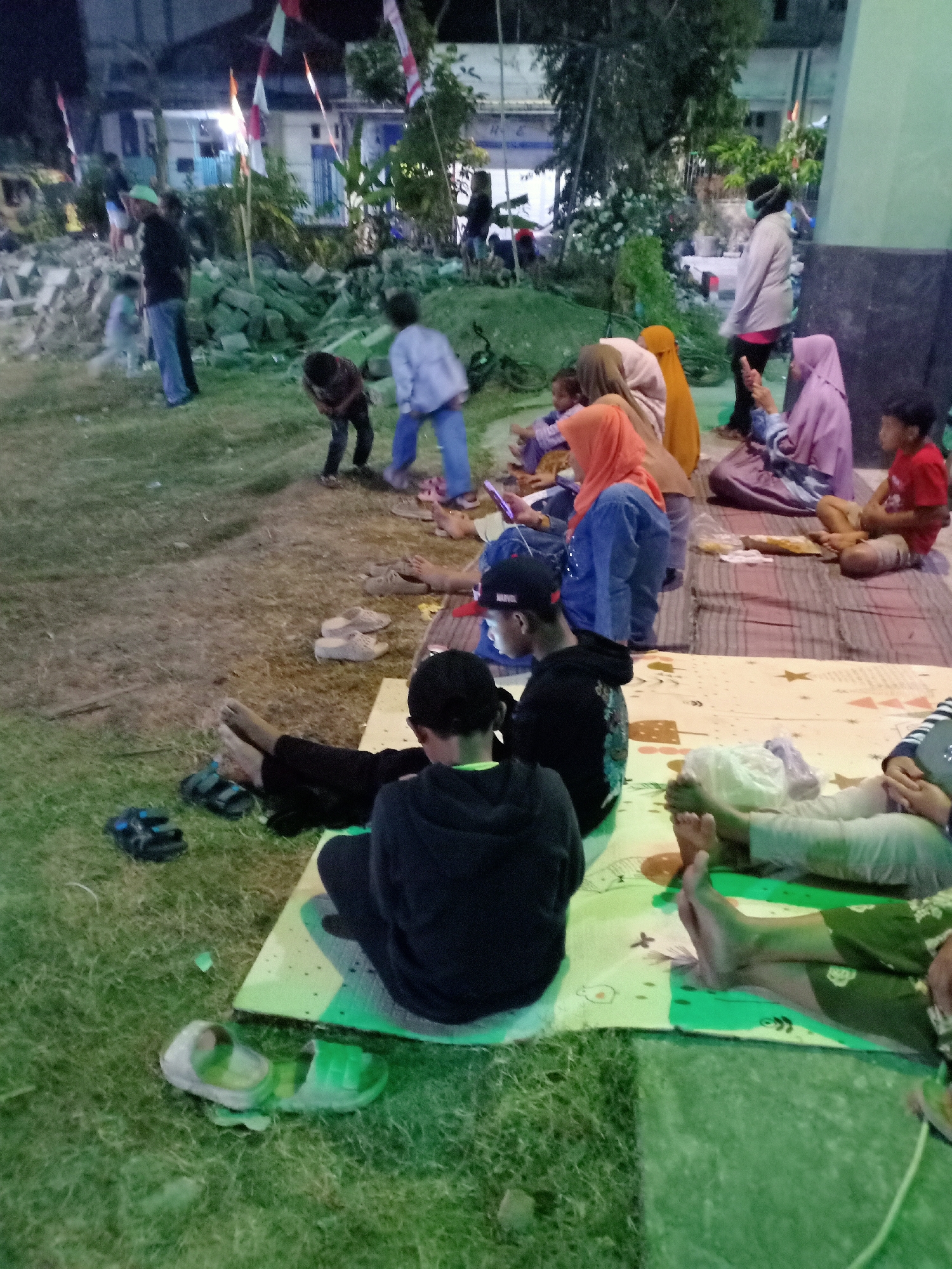 In Sumber Tlaseh, Indonesia, a group of people are gathered outdoors, possibly for an event or gathering, during the evening. The ground is a mixture of grass and dirt, with some areas covered by mats. The setting appears to be near buildings, some of which are illuminated.
In the foreground, several individuals are seated or reclining on mats. One person in a black hoodie is facing away from the camera. To their right, another person's legs are visible, wearing green patterned pants. Further to the right, two more individuals are seated on mats, their legs extended. Scattered footwear, including sandals, are present on the grass and near the mats.
In the midground, a group of children and adults are also seated on mats. Some of the adults are wearing headscarves. A child in a blue shirt is visible playing, while another child in a dark shirt appears to be bending over. Several pairs of sandals are also placed on the ground.
In the background, several people are standing and walking. One person in a light-colored jacket and dark pants is walking away from the camera. Other individuals are less discernible due to the distance and lighting. Decorations, possibly flags, are visible strung between poles, suggesting a celebratory or communal event. Debris or construction materials are present in the far left background, indicating potential ongoing work or a past event. The overall atmosphere seems relaxed, with people engaged in various activities.