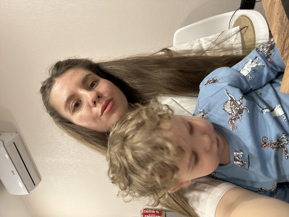 An indoor scene in the city of Shipulino, Russia, features an adult female and a young child. The female, with long light brown hair and a neutral expression, is looking directly towards the camera, positioned slightly to the right of the frame's center. She is wearing a white collared garment. A child with curly light brown hair is seated in front of the female, looking downwards and slightly left. The child is wearing a light blue long-sleeved shirt patterned with various dinosaurs. In the upper left background, a white wall-mounted air conditioning unit is visible. To the adult's left, a red and white "Choco-Pie" box is partially discernible on a surface. To the right, a white chair with a circular, light brown woven seat cushion is present. The background wall is a plain, light color with faint vertical texture. A wooden surface is partially visible in the lower right foreground.