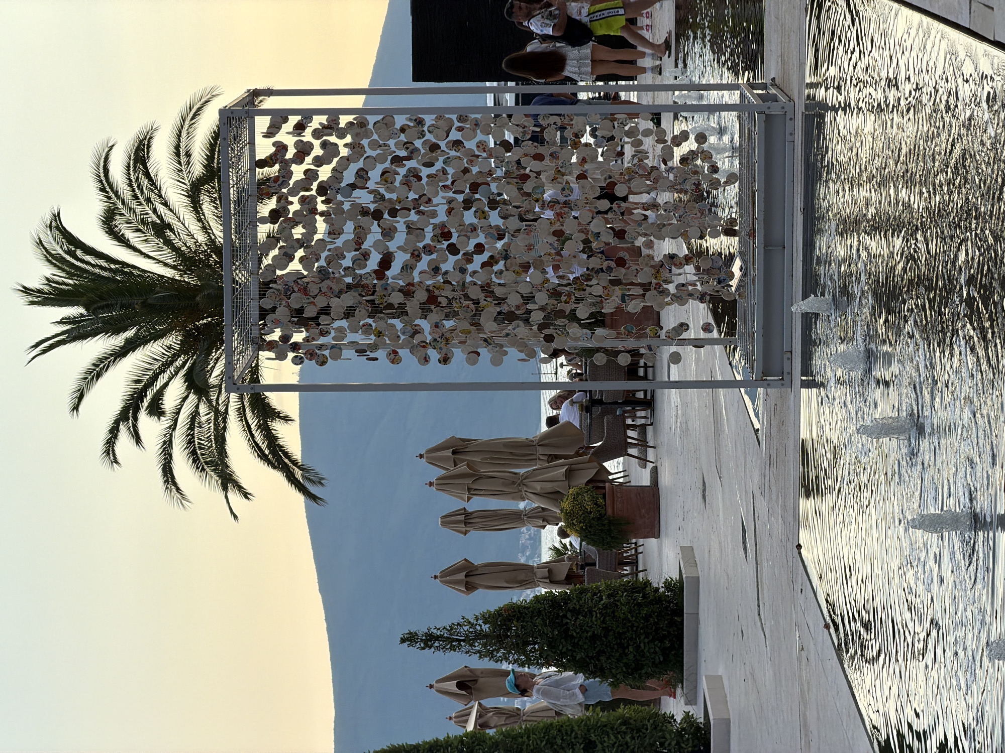 This media file depicts an outdoor scene in Tivat, Montenegro, likely during the late afternoon or early evening. The setting appears to be a waterfront area with a fountain or water feature. In the foreground, a tall palm tree is visible to the left, with its fronds reaching towards the sky. To the right of the palm tree is a large, open-sided metallic cube structure adorned with numerous small, circular decorative elements.
The background reveals a paved area with tables and chairs, suggesting a restaurant or cafe. Several closed beige umbrellas are arranged neatly in front of the seating area. Beyond the paved area, a body of water is visible, reflecting the sky and surrounding structures. In the far background, a mountainous landscape meets the horizon, with the sky transitioning from a soft yellow or pale orange near the horizon to a pale blue or grey higher up, indicating sunset or sunrise.
Several people are present in the scene, some walking and others seated at the tables. Their attire suggests a casual, warm-weather environment. One person is visible near the fountain, and others are seated further back. The overall atmosphere appears relaxed and serene. The water in the fountain is rippling, with visible jets of water. The scene is captured in a vertical orientation. There is no visible text in the media file.